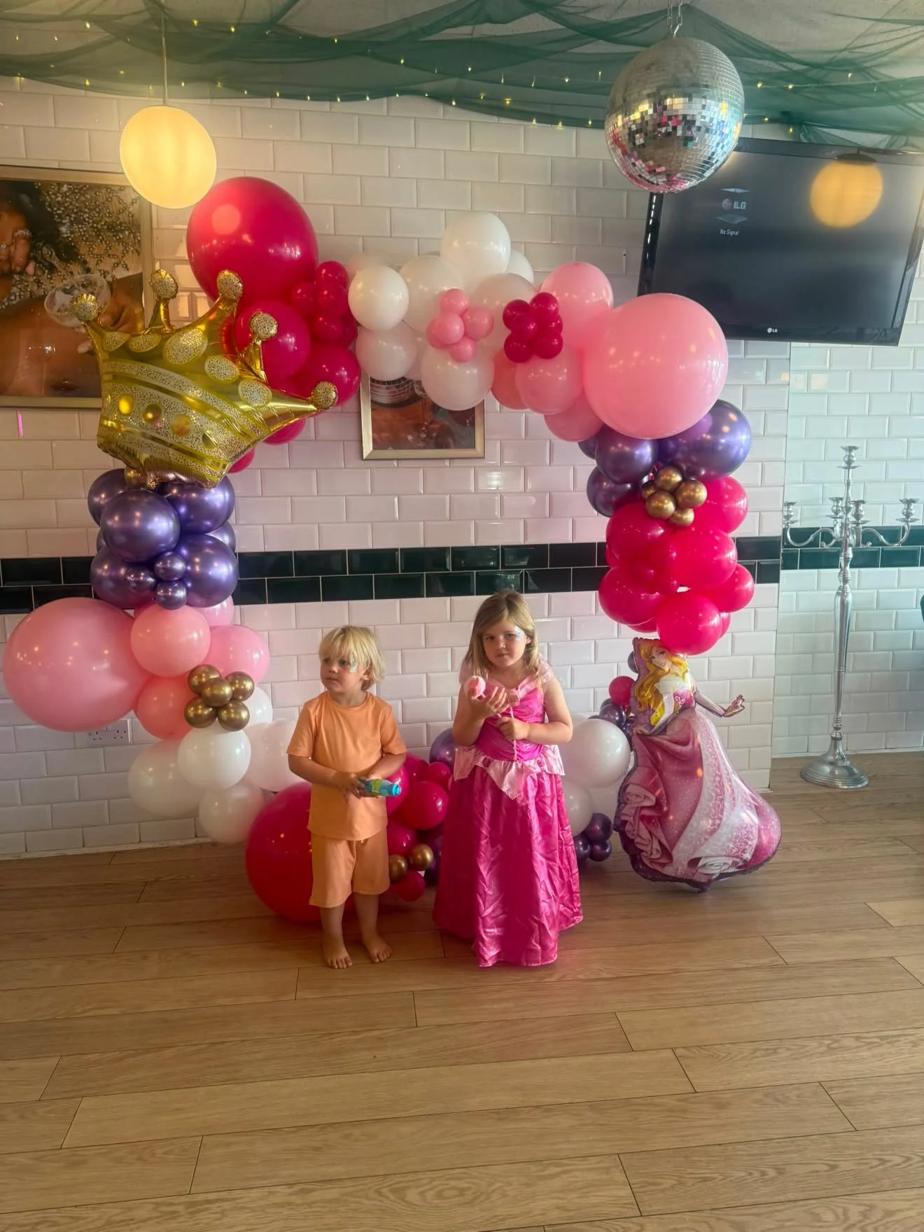 Children at a birthday party standing in front of a balloon arch with pink, white, purple, and gold balloons, a golden crown balloon, and a Disney princess balloon. The room has a candelabra, a wall-mounted TV, and framed pictures on a white brick wa