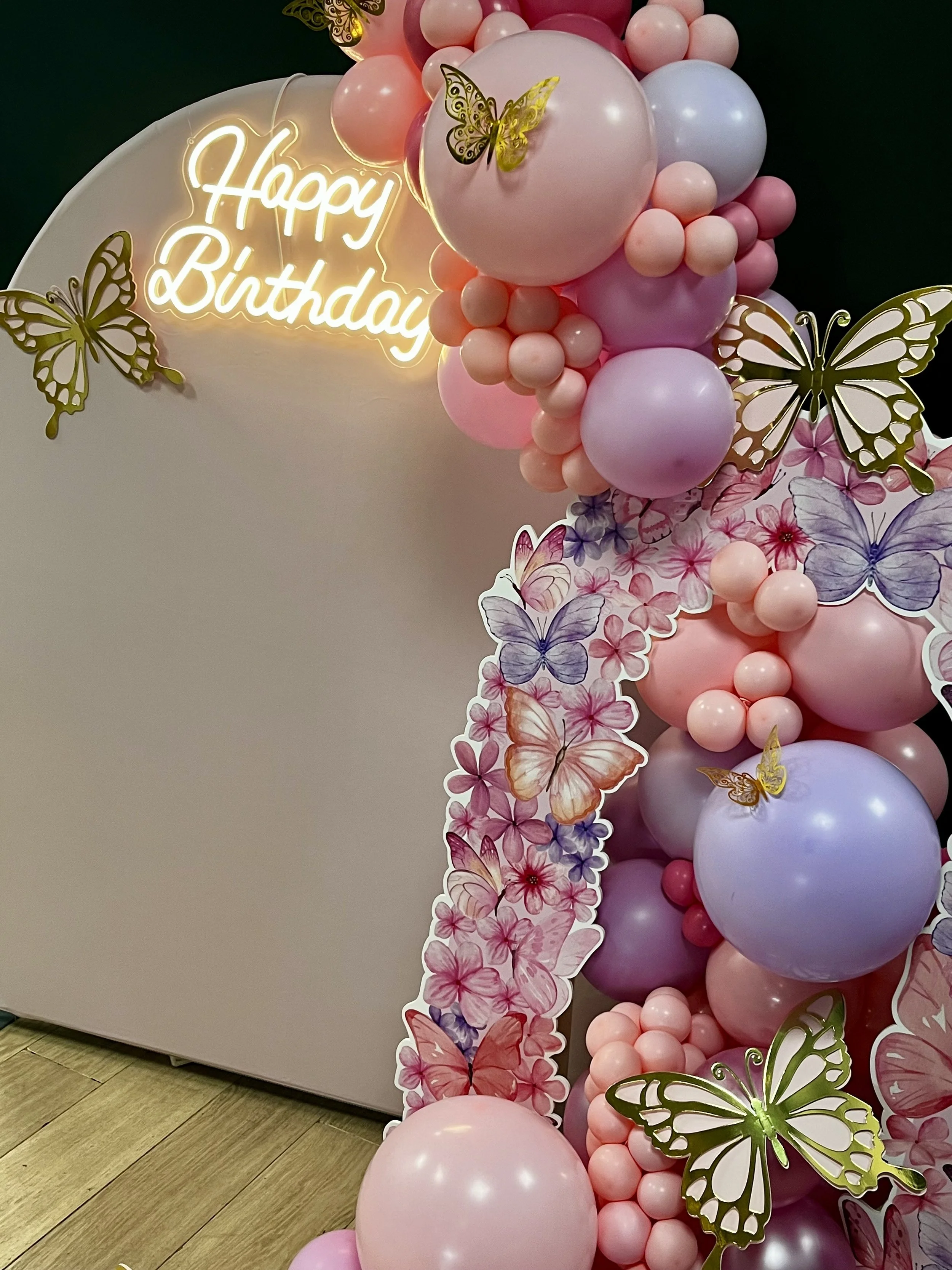 Colorful balloon arrangement in pink, purple, and white with butterfly decorations and a neon sign that reads 'Happy Birthday'.