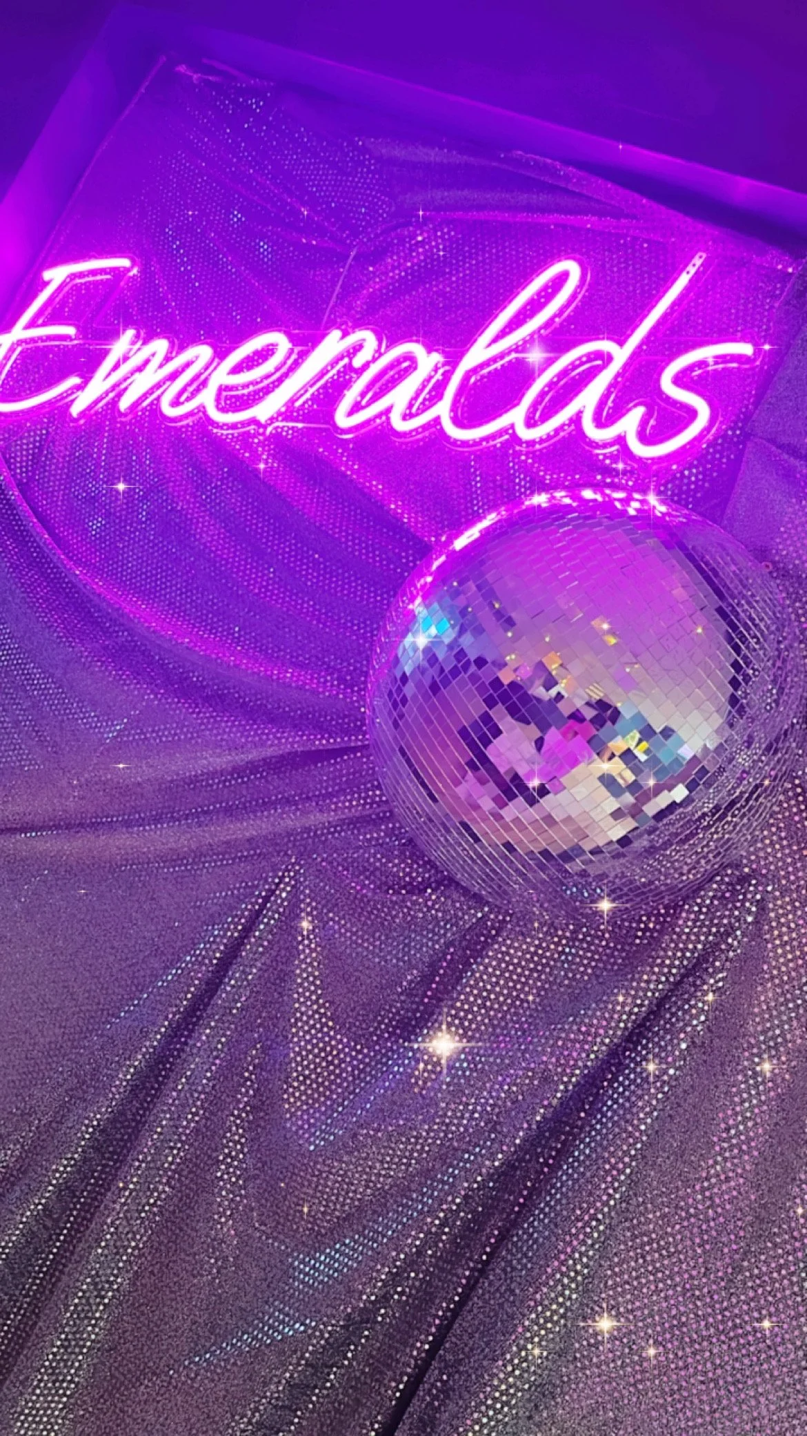 A neon purple sign that says 'Emeralds' in cursive, alongside a disco ball on a dotted purple backdrop.