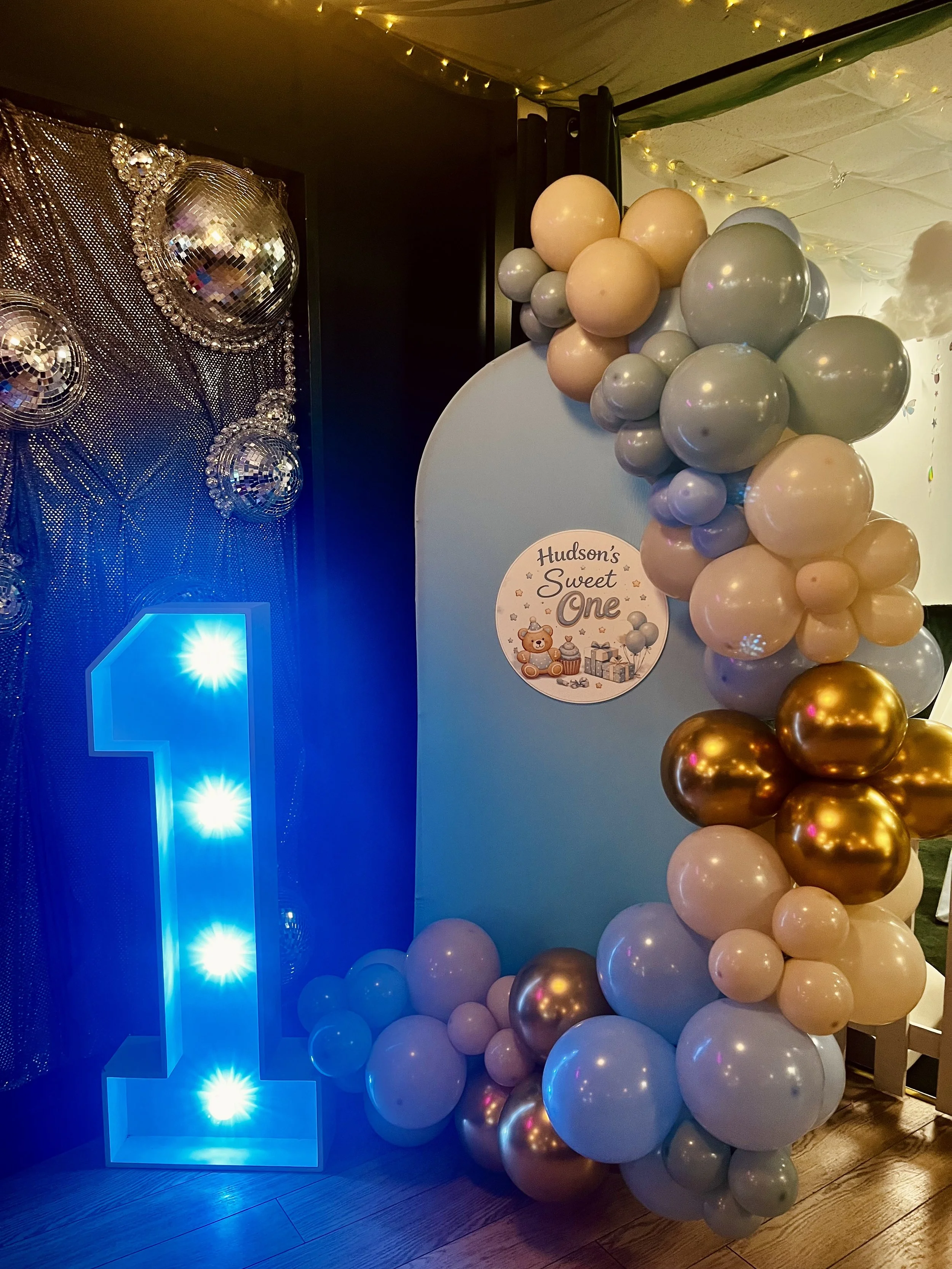 Decorative blue illuminated block next to a cluster of pastel and metallic balloons, with a sign reading 'Hudson's first birthday' and a teddy bear holding a cupcake surrounded by presents, at a birthday celebration.