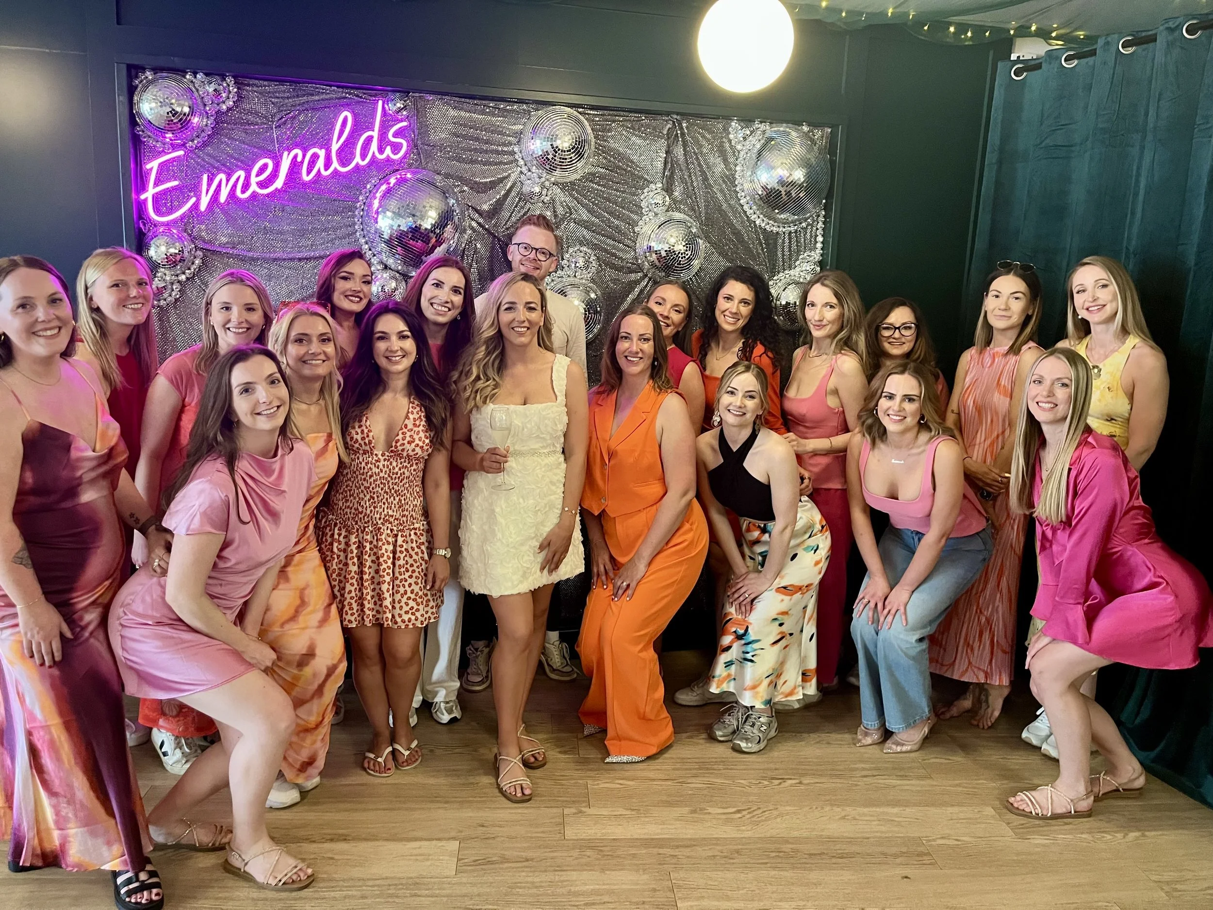 A group of women and one man gathered together in front of a silver, disco-ball decorated backdrop with a neon sign that says "Emeralds." They are smiling, dressed in colorful, summery outfits, and appear to be at a celebration or party.