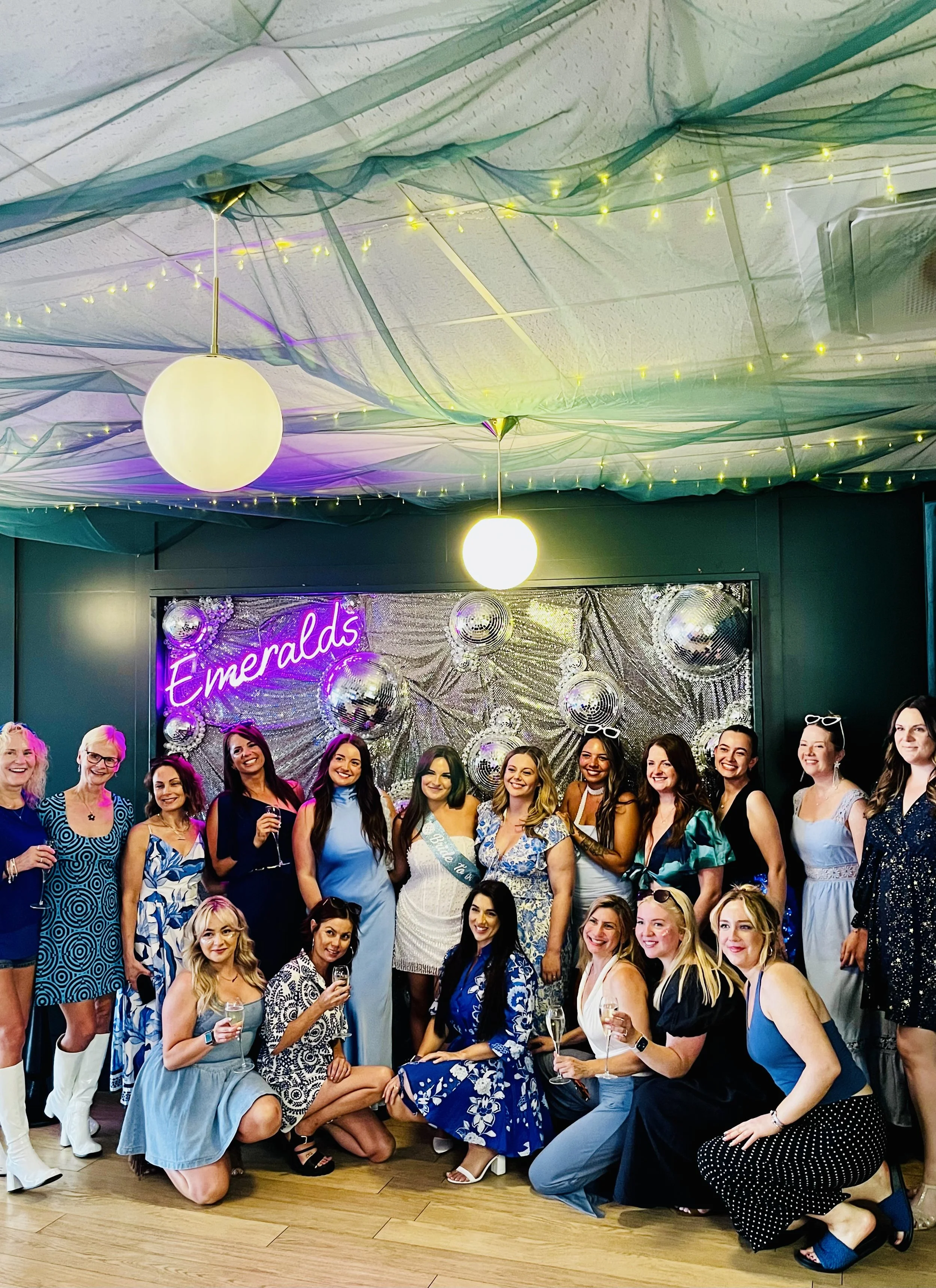 Group of women at a celebration, dressed in elegant outfits, holding drinks, with a decorated background and a neon sign that says 'Emeralds'.