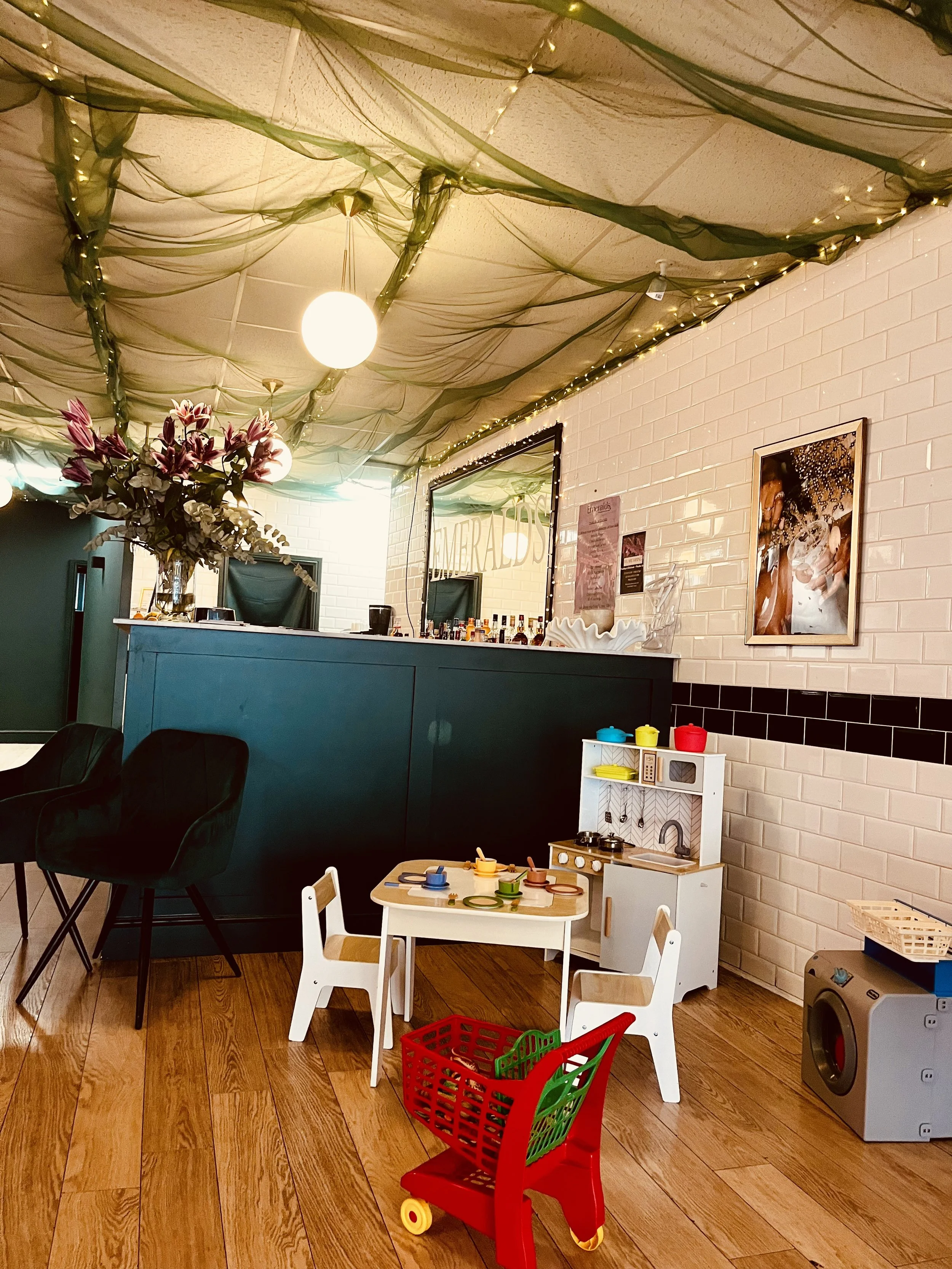 A children's play area with a small white table and chairs, a red shopping cart toy, a toy kitchen set, and toys on the wooden floor. In the background, there are black chairs, a bar area with bottles, framed pictures on the white brick wall, and dec