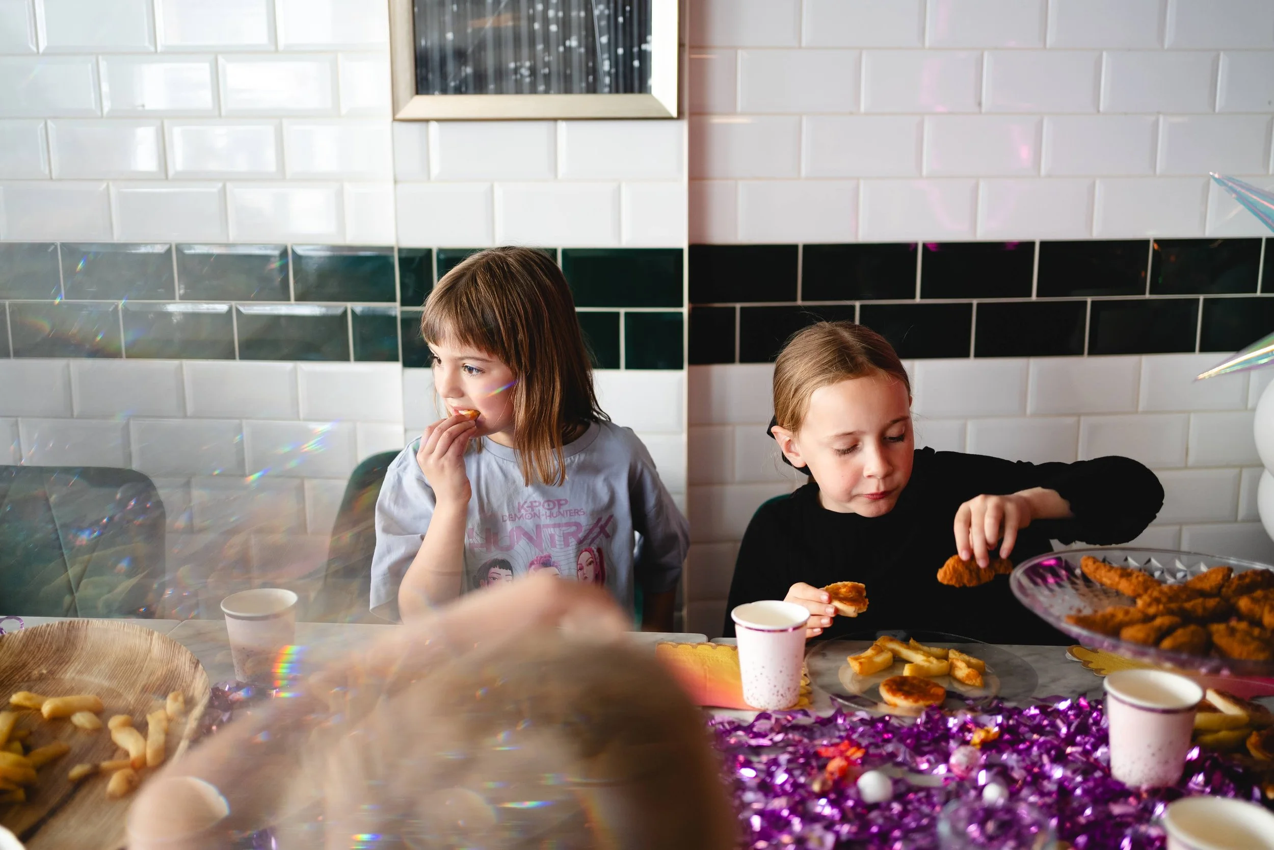Two children sitting at a table enjoying food at a celebration, with a white tiled wall and a window in the background, decorated with purple confetti and party dishes at emeralds chelmsford