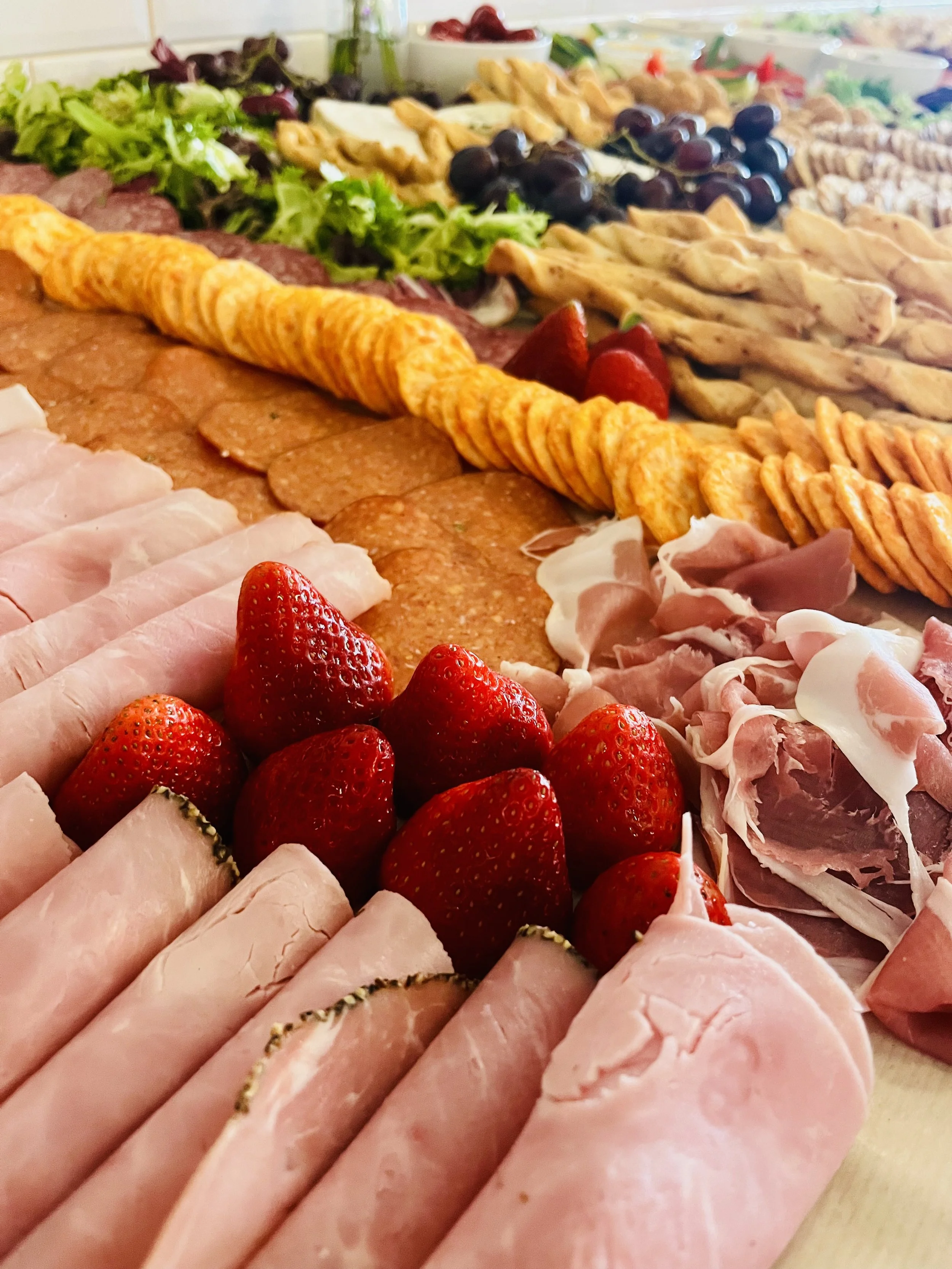 A platter of sliced ham, fresh strawberries, slices of turkey, salami, and various cheeses, with a variety of fresh vegetables and other snacks in the background.