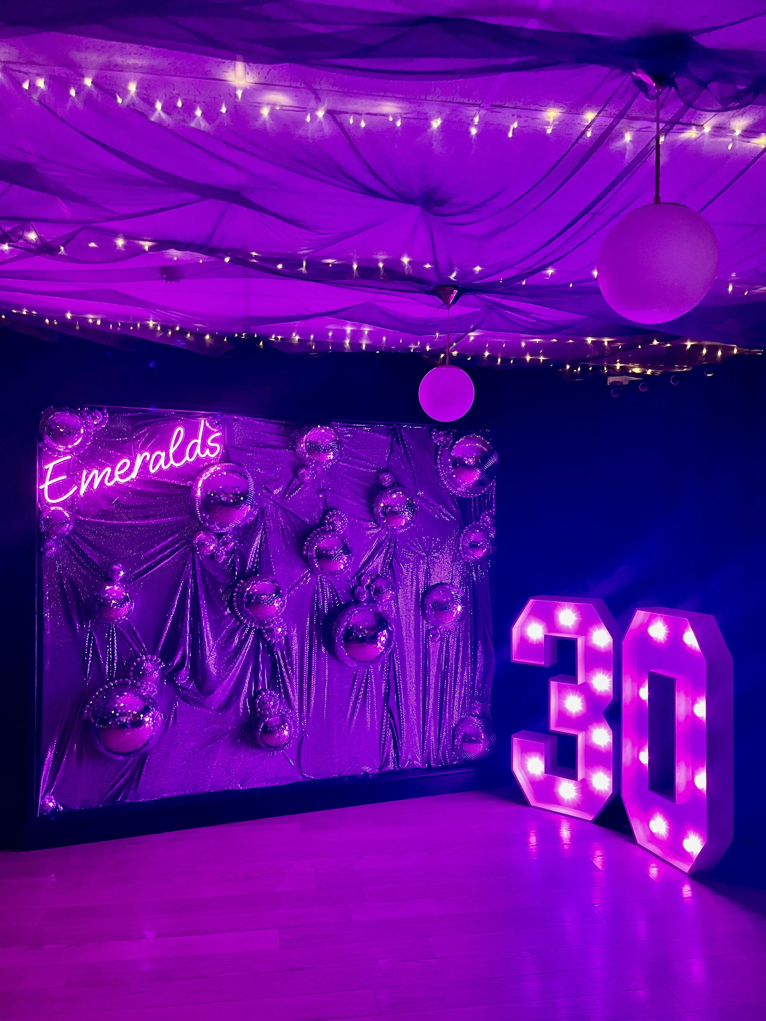 Decorative party setup with purple lighting, a backdrop labeled 'Emeralds' with metallic balloons, and large illuminated numbers '20' in pink light.