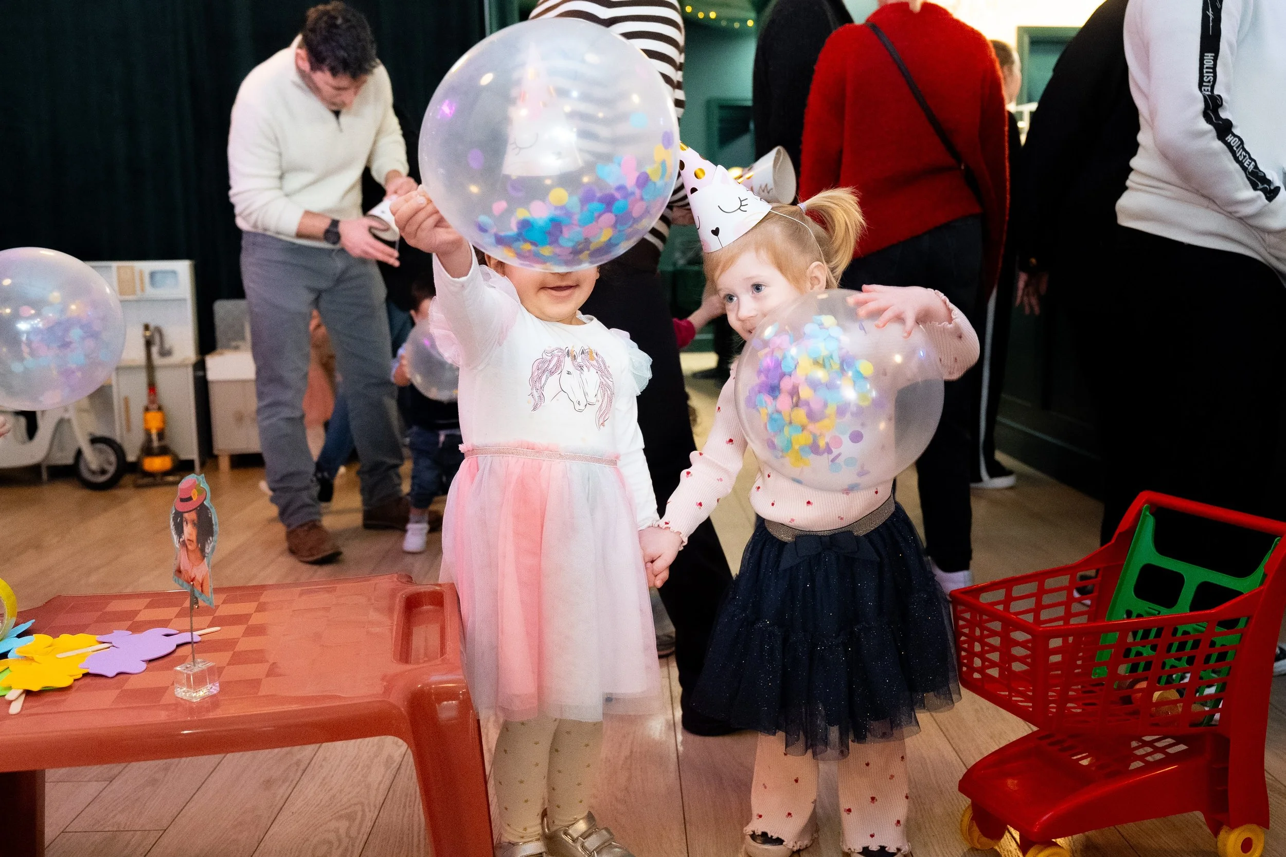 Two young girls are celebrating a birthday with party hats. One girl has a unicorn shirt and a pink skirt, while the other girl wears a pink sweater with red hearts and a dark blue skirt. They are holding hands and each has a clear balloon filled wit