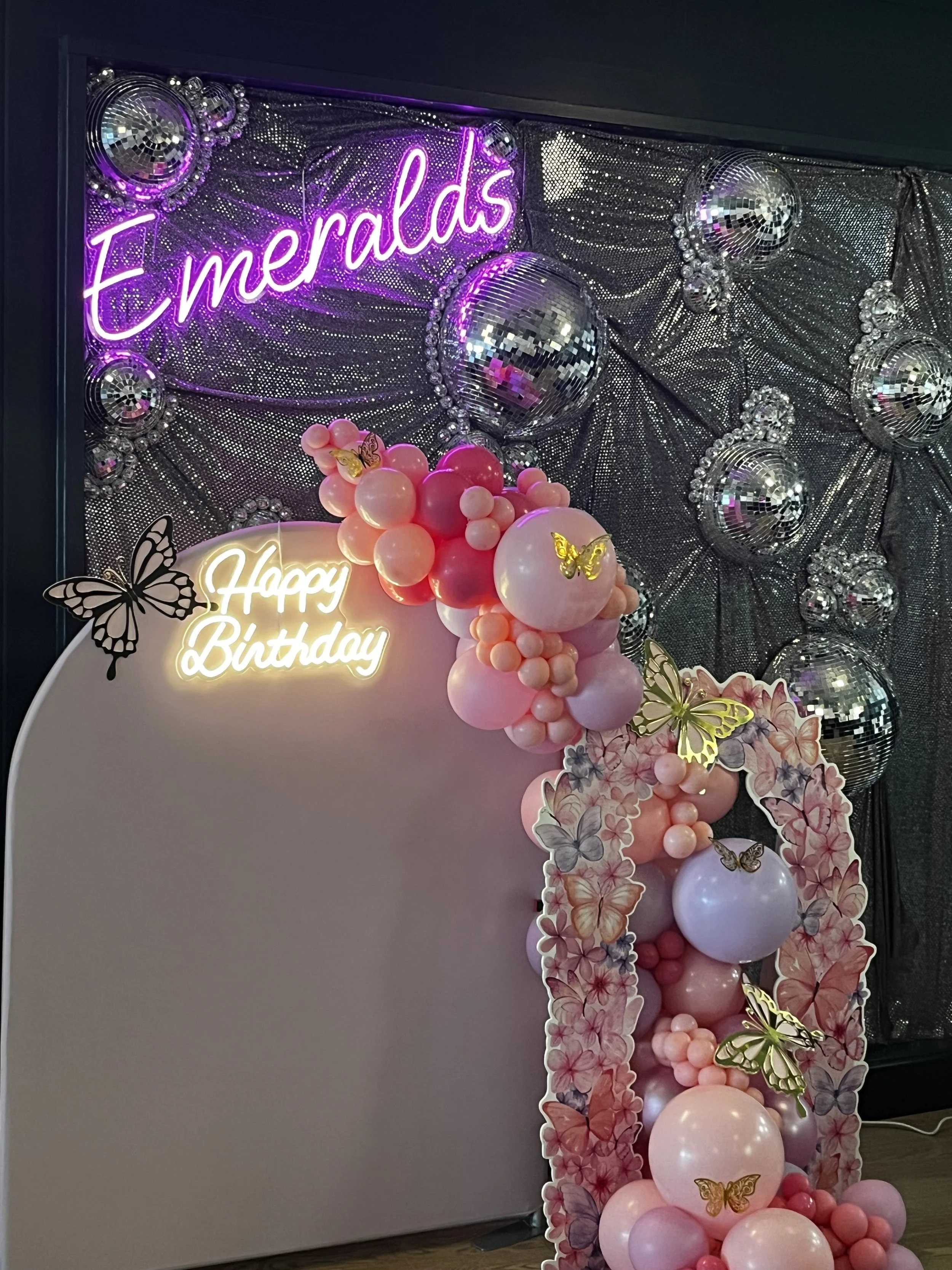 Decorative birthday display with pink and white balloons, butterfly cutouts, a pink 'Emeralds' neon sign, and a pink and white 'Happy Birthday' sign, set against a silver sequin backdrop with disco balls.