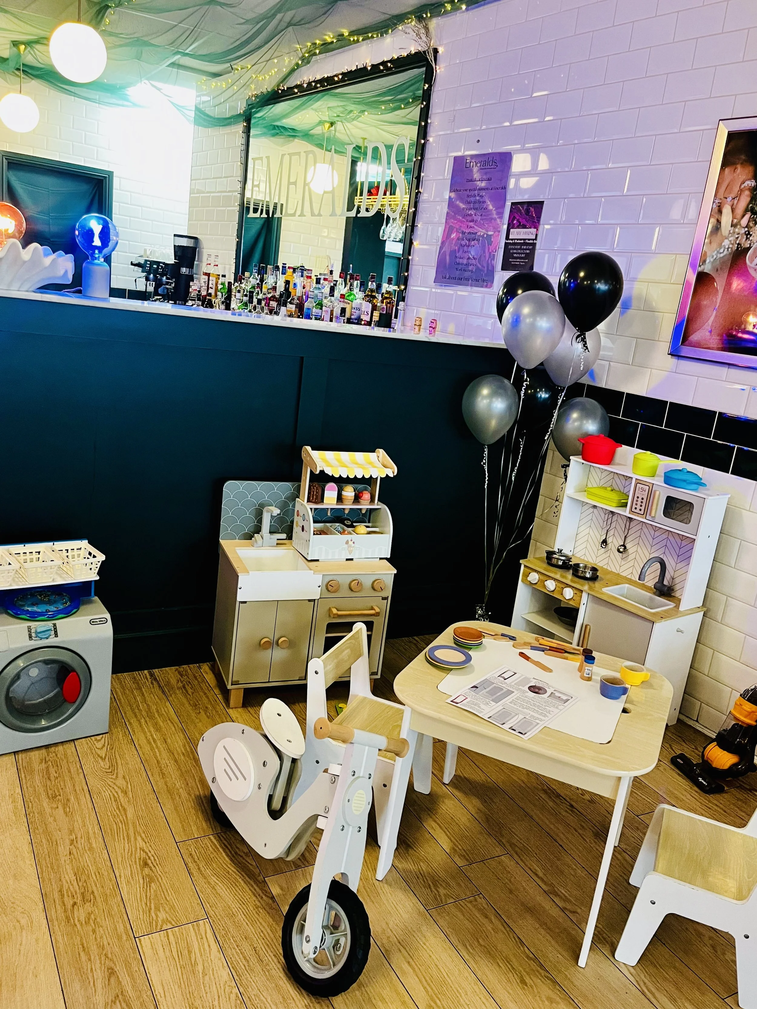 A children's play area with miniature furniture, including a white table with chairs, a toy kitchen, and a doll ride-on quad. There are balloons in black, silver, and gray. The background features a bar setup with bottles, a mirror with a sign readin