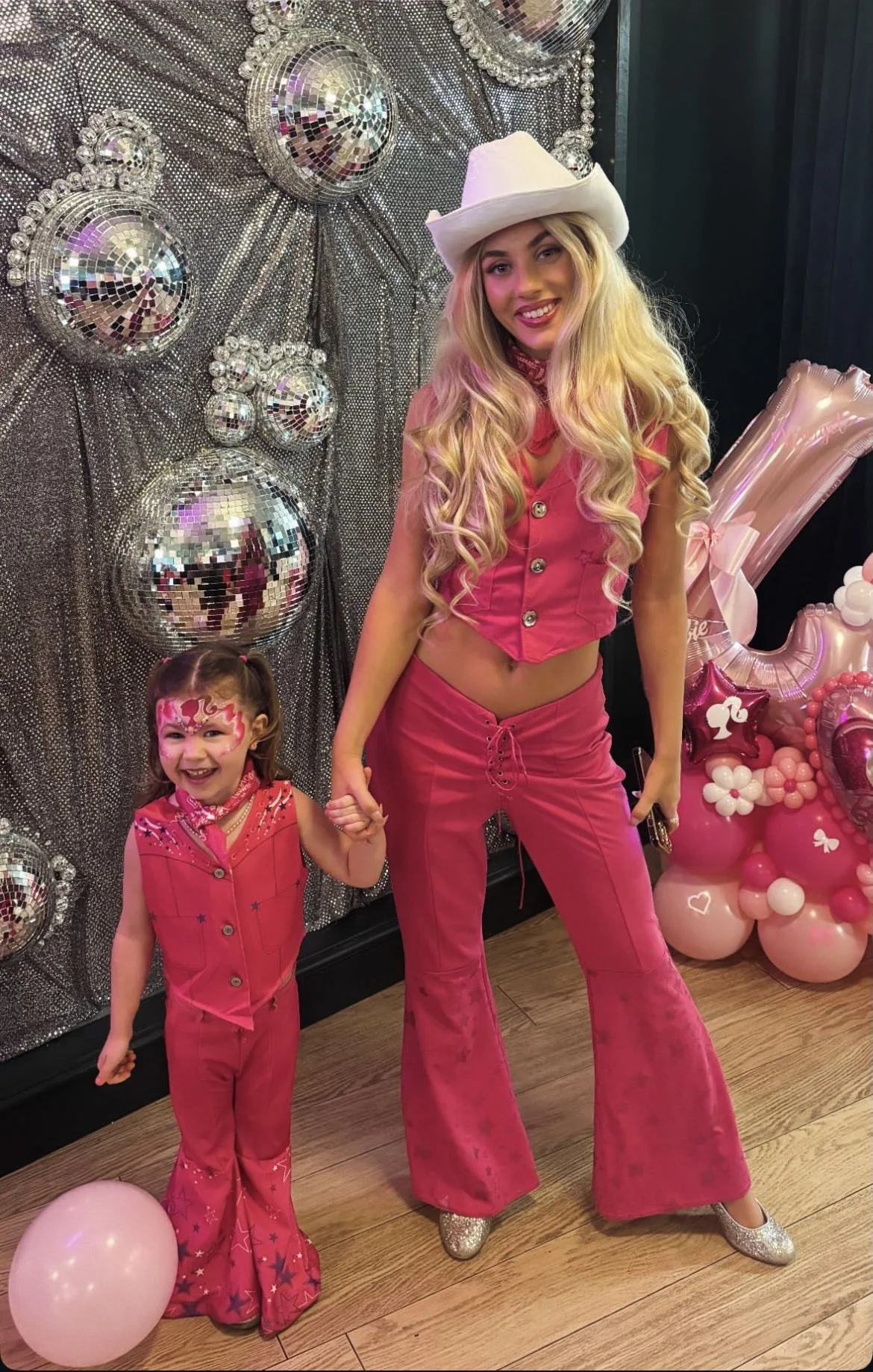 A woman and a young girl dressed in matching pink outfits, holding hands at a festive party. The woman is wearing a white cowboy hat and pink bell-bottom pants, while the girl has face paint and a sleeveless pink vest with star patterns. The backgrou