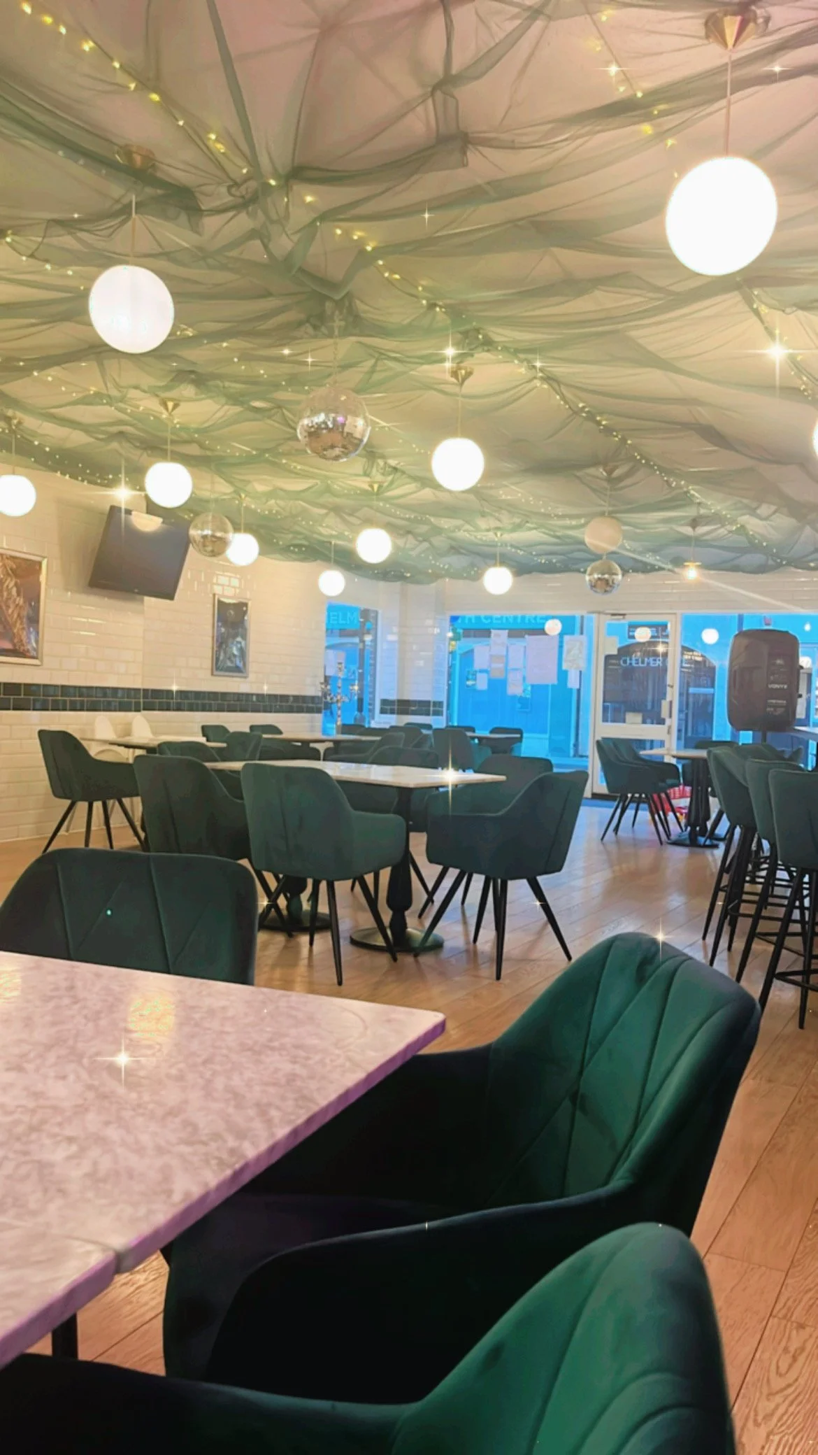 An interior view of a restaurant or dining area with tables and green cushioned chairs. The ceiling is decorated with draped fabric, string lights, illuminated globe pendant lights, and small disco balls. Large windows at the front allow natural ligh