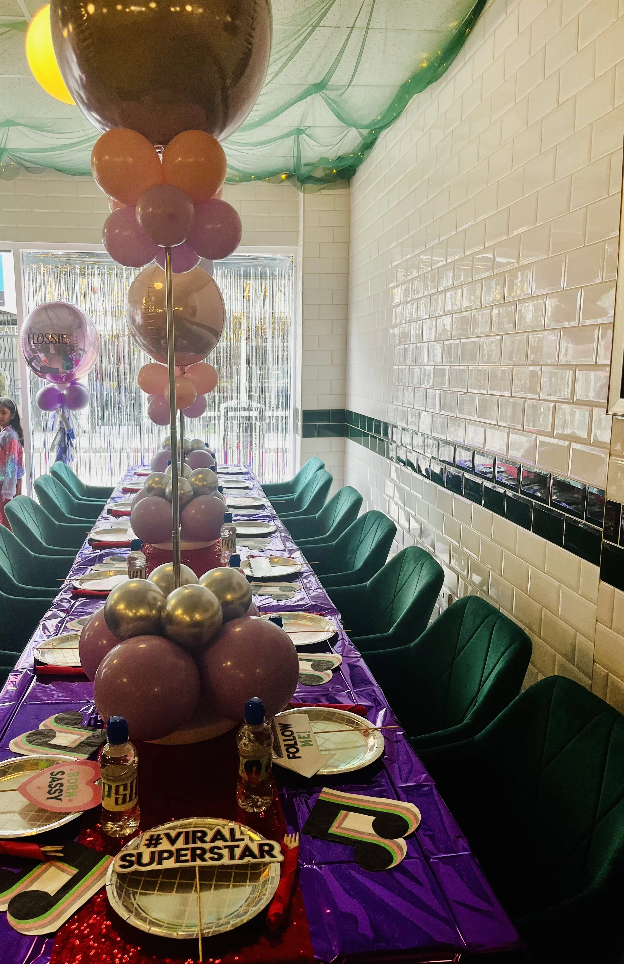 Party table decorated with purple and red tablecloths, colorful balloons, and themed plates, with a long row of chairs and a balloon centerpiece in a decorated party room.