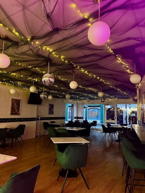 Empty event space with tables and chairs, decorated with hanging white spherical lights and strings of yellow fairy lights on the ceiling. Disco balls are also hanging from the ceiling. Large windows at the front provide a view outside.