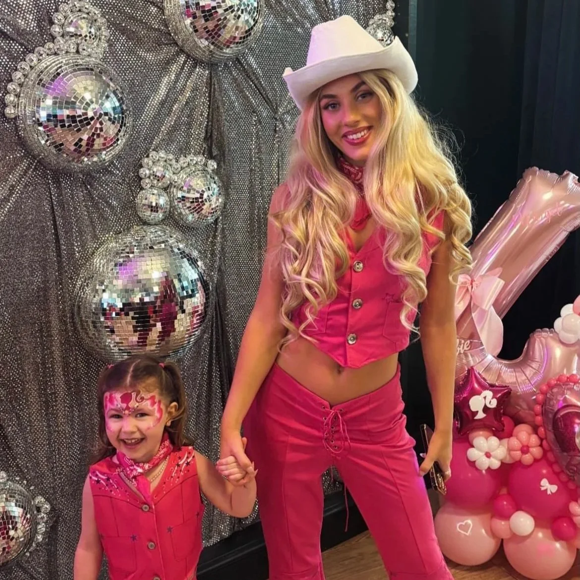 A young girl and a woman dressed in pink at a party, holding hands. The girl has face paint and a pink vest, and the woman is wearing a cowboy hat and pink outfit. They are standing in front of a glittery backdrop with disco balls and pink balloons, some shaped like hearts and stars.