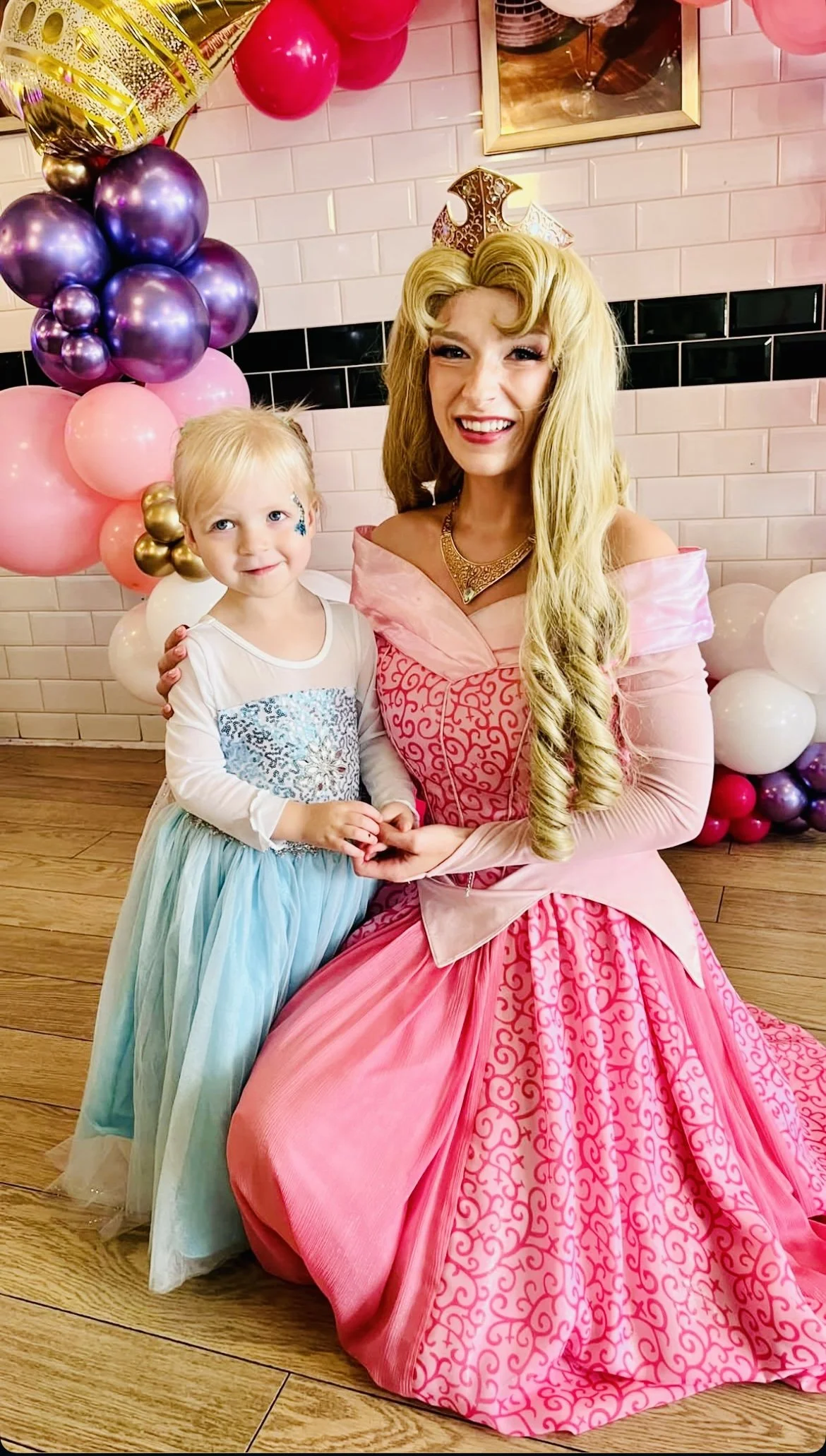 A woman dressed as Princess Aurora from Disney's Sleeping Beauty, sitting next to a young girl with face paint, at a birthday party with pink, purple, white, and gold balloons and decorations. kids celebration at emeralds chelmsford