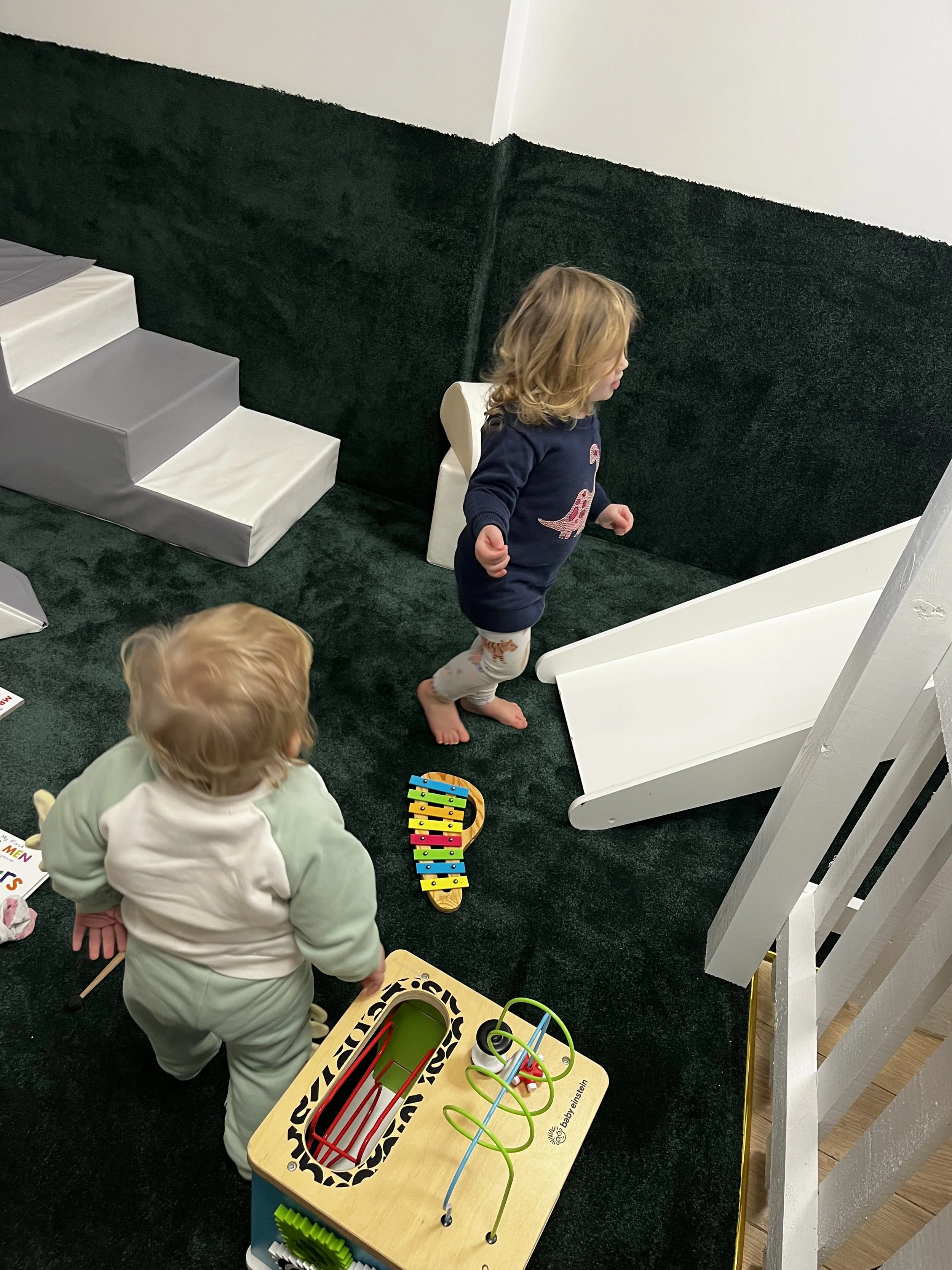 Children playing with toys in a playroom with black carpet, white stairs, a xylophone, and a white slide.