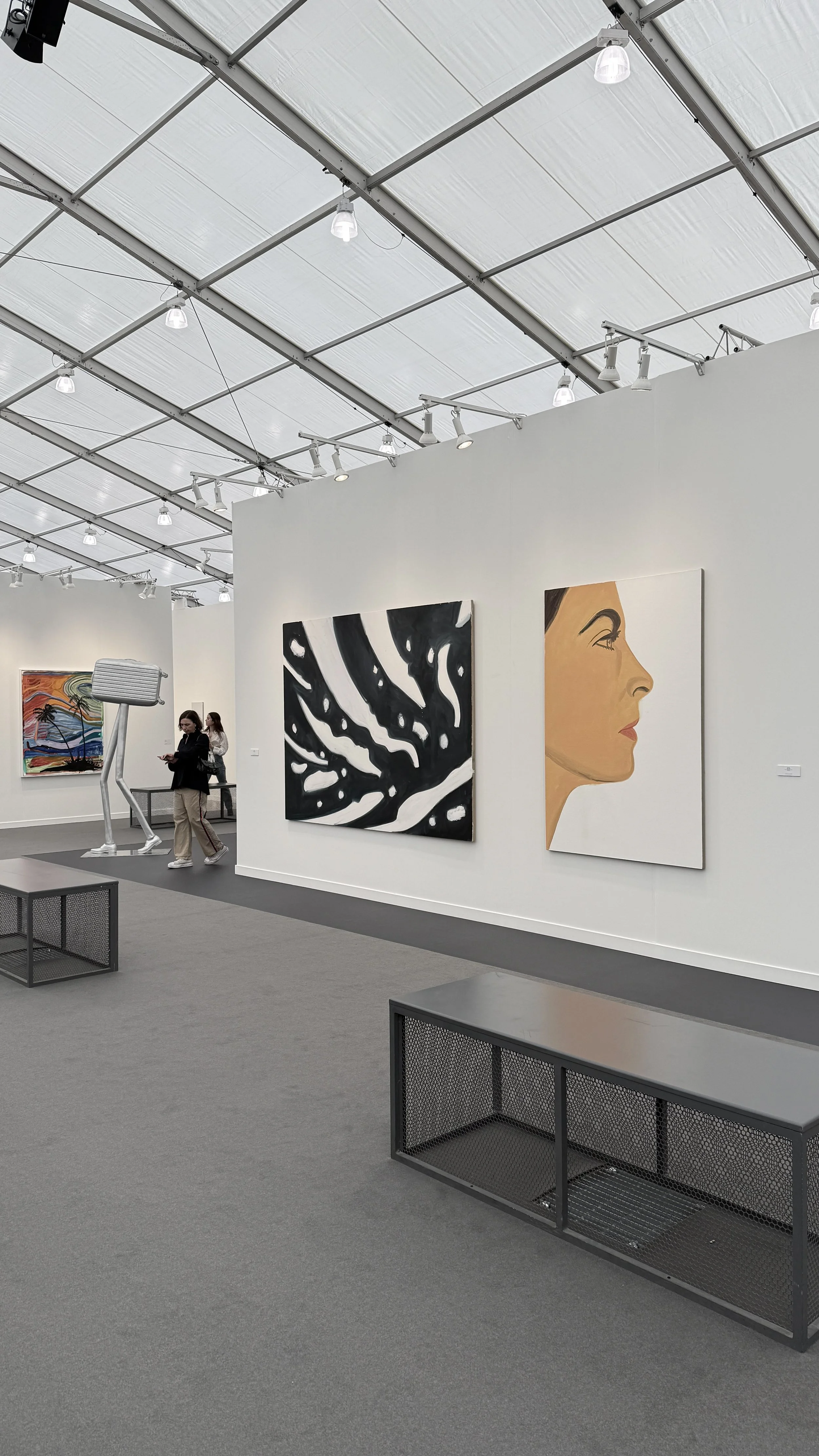An art gallery with white walls displaying various paintings, including a colorful landscape, a black-and-white abstract piece, and a profile of a woman's face. There are a few visitors and industrial-style lighting fixtures.