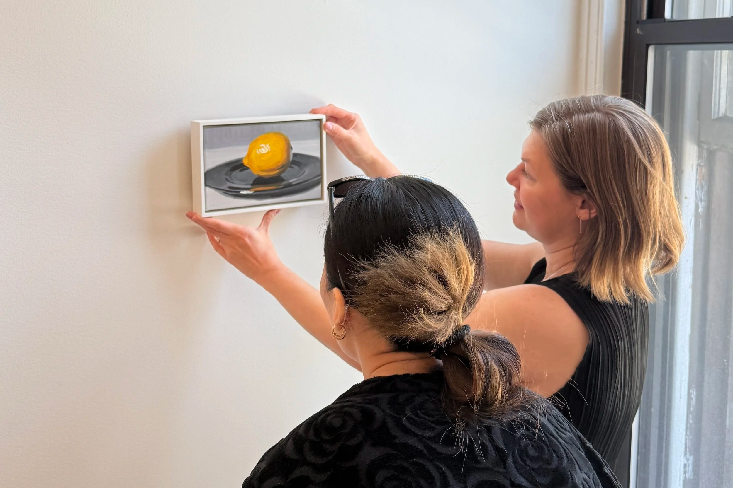 Two women are looking at a painting of a lemon by Cynthia Daignault on a white wall in the gallery.