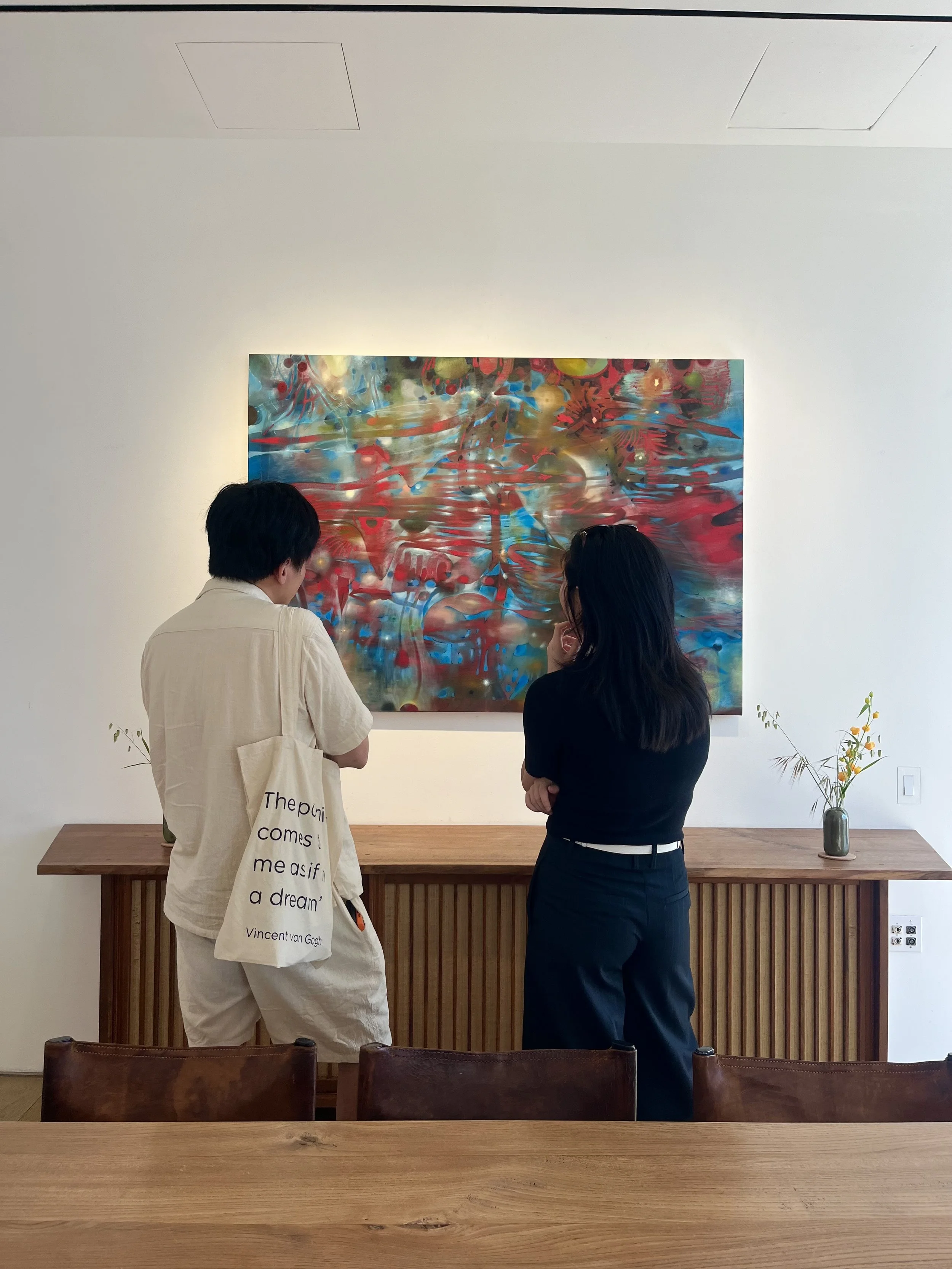 Two people standing in front of an abstract colorful painting in a gallery, with a wooden table and chairs in the foreground and a white wall with ceiling lights in the background.