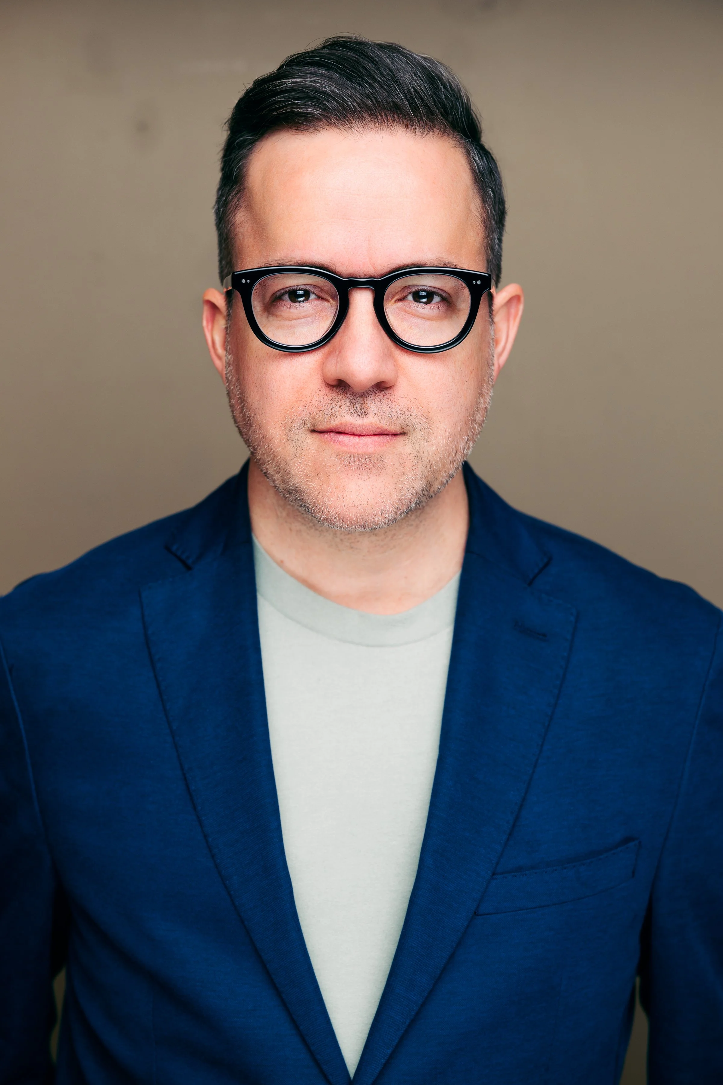 Close-up of a man with dark hair, wearing black glasses, a light grey shirt, and a navy blazer, posing against a plain beige background.