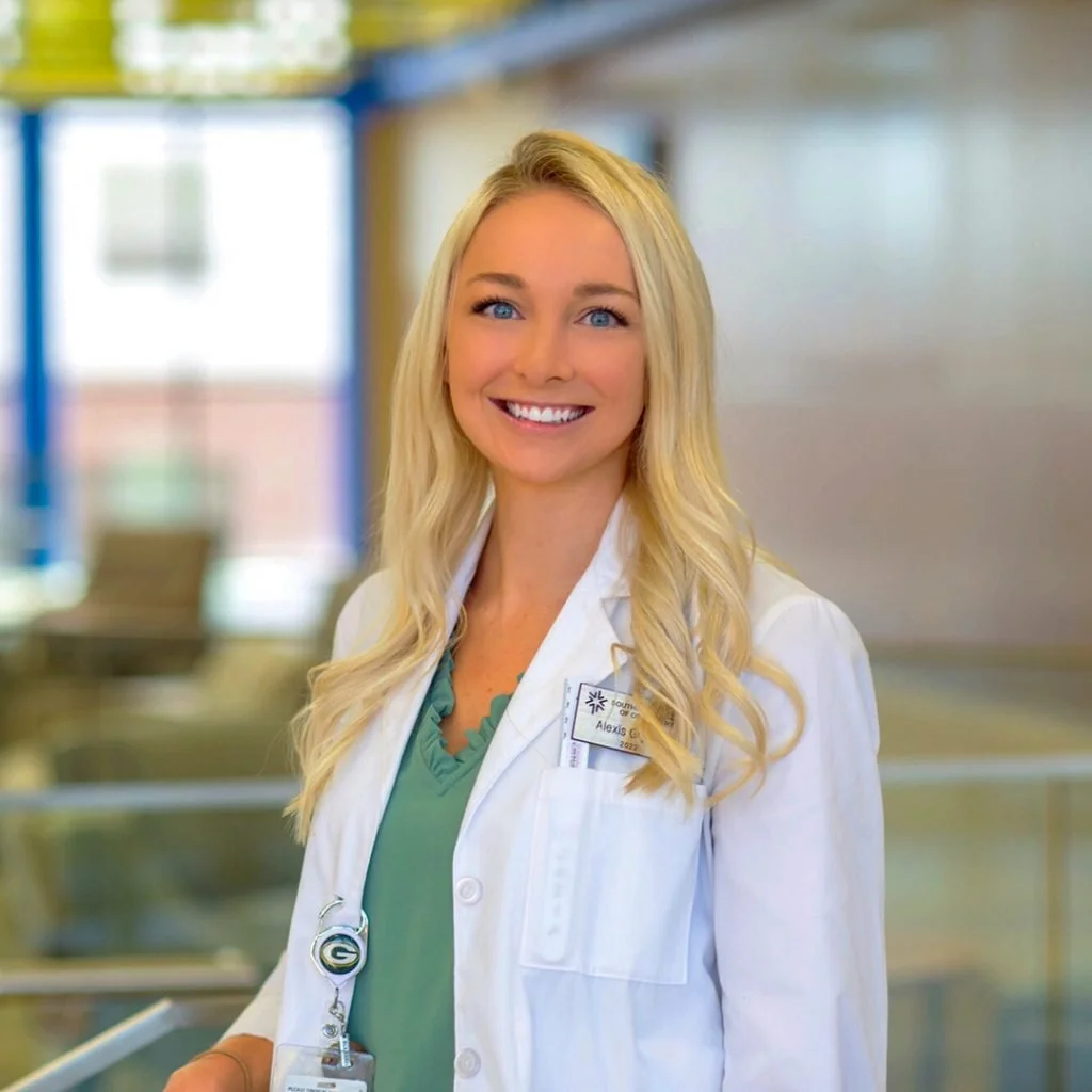 A smiling female healthcare professional with blonde hair, wearing a white coat and green scrubs, standing in a medical facility.