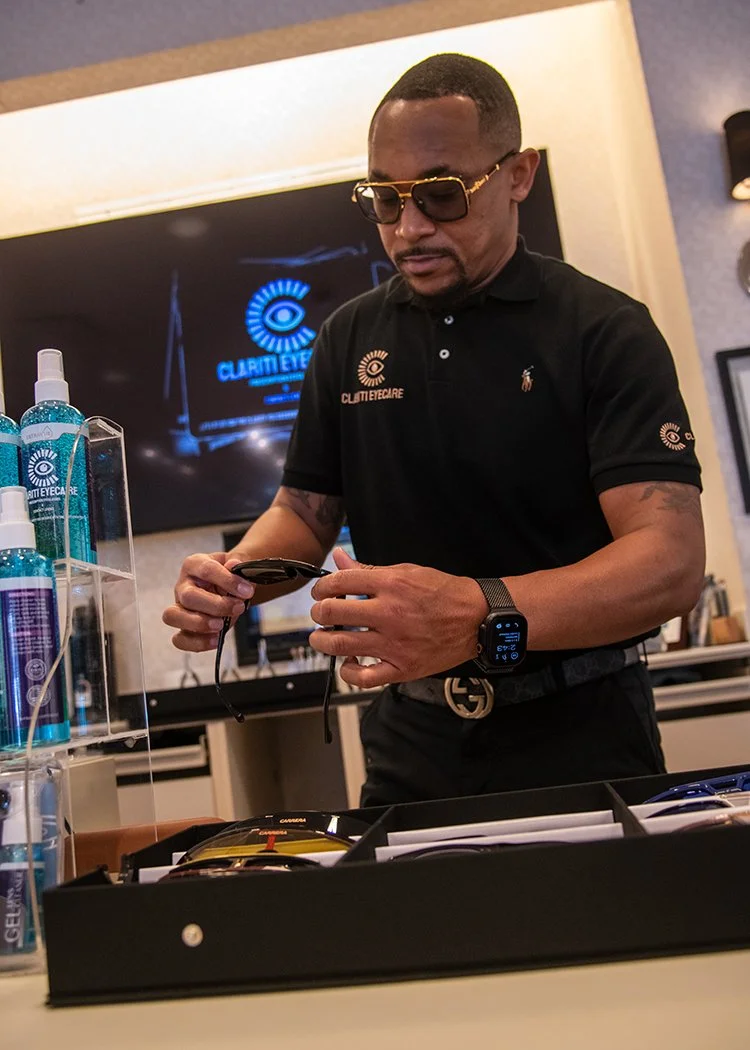 A man in sunglasses and a black polo shirt with the logo 'Clutti Eyecare' is standing at a table, holding a pair of glasses. There are bottles of eyewear cleaning solution on a display shelf nearby and a monitor in the background displaying the same logo.