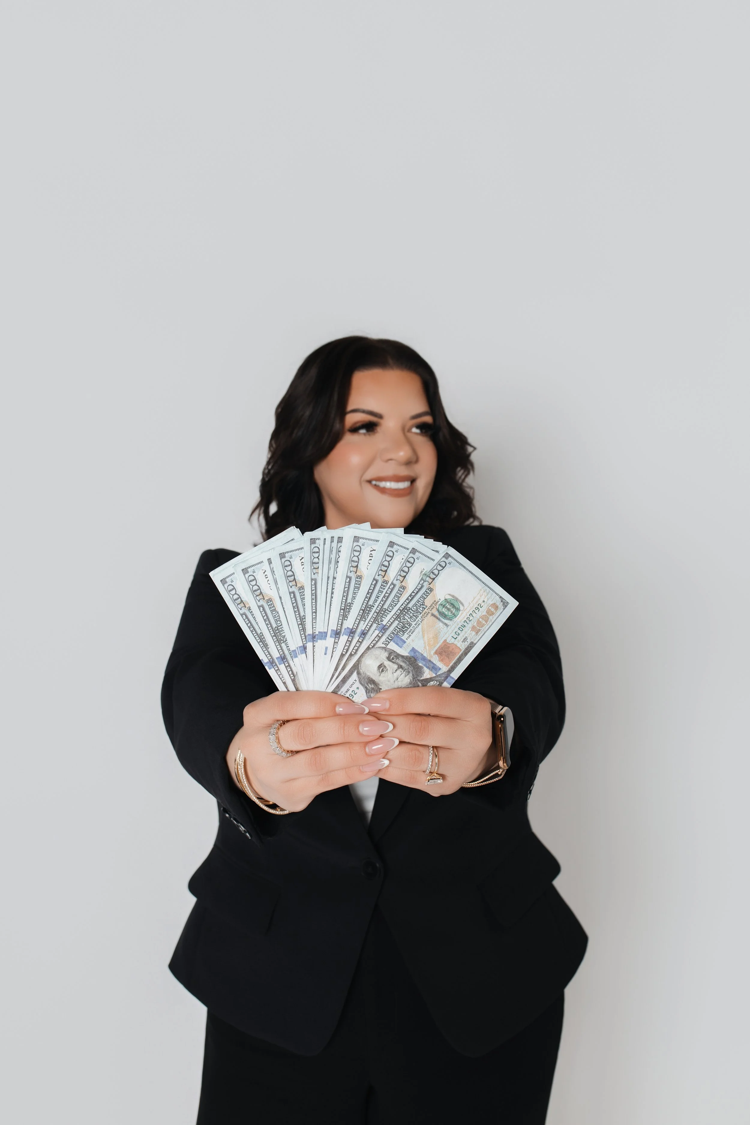 A woman with dark wavy hair, wearing a black suit, holding a fan of U.S. hundred-dollar bills and smiling against a plain gray background.