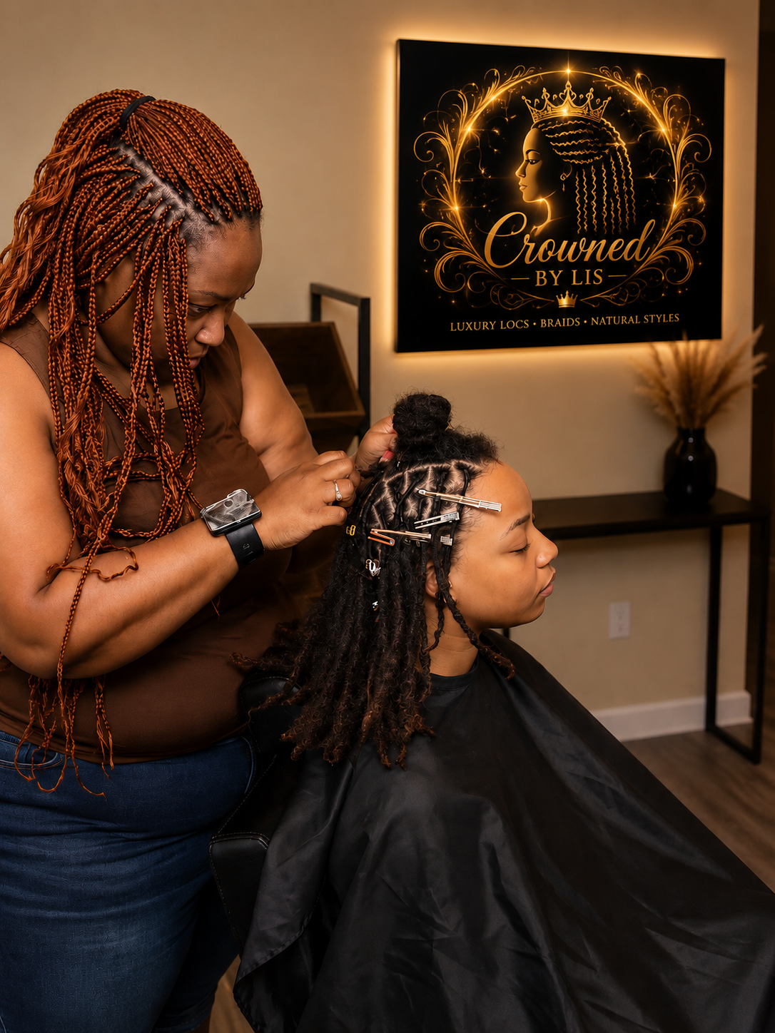 A woman getting her hair styled in braids at a salon, with a stylist working on her hair, in front of a sign that reads 'Crowned by Lis' and advertises luxury locs, braids, and natural styles.