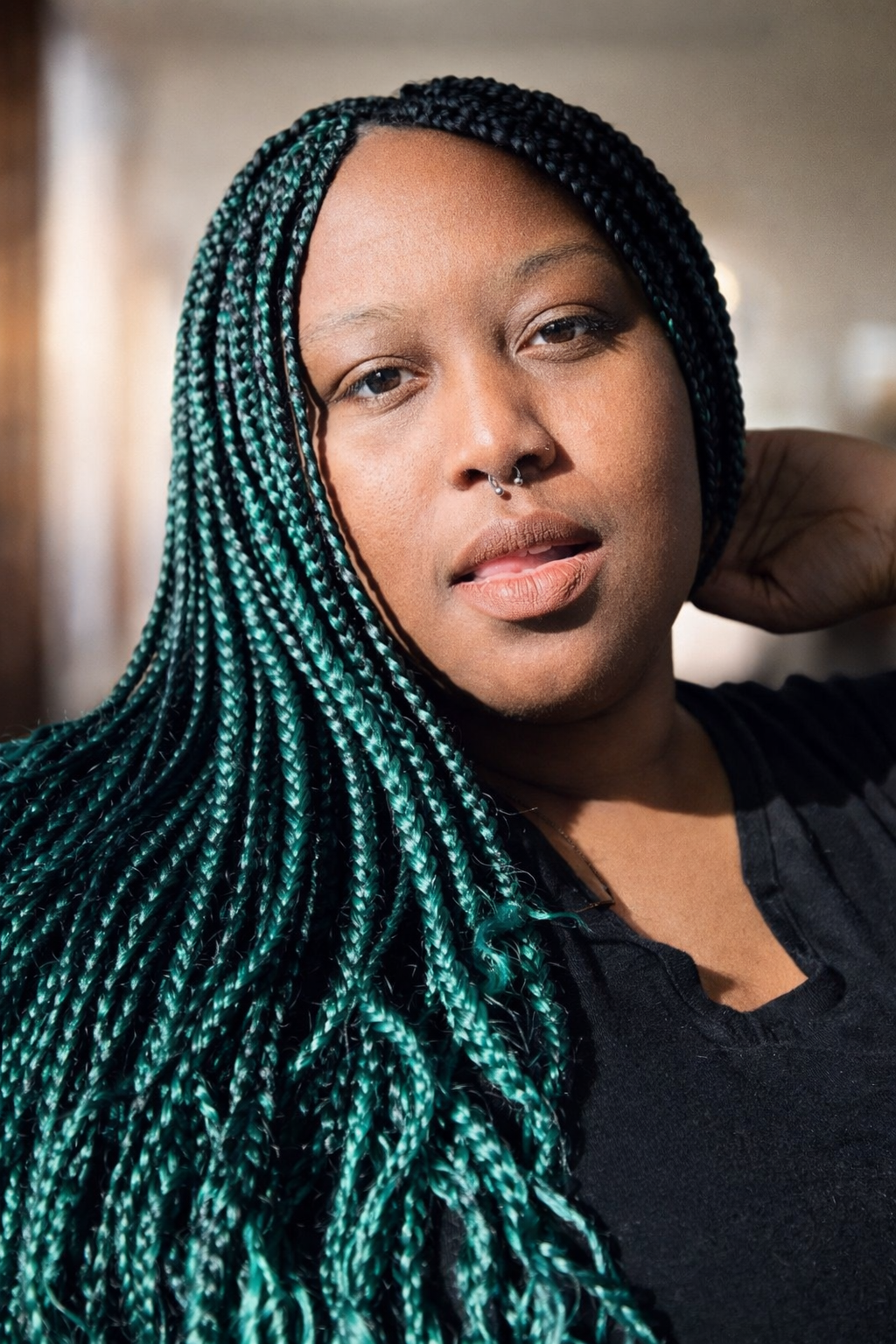 Close-up portrait of a woman with braided hair in shades of teal and black, posing with her hand behind her neck in a softly lit indoor setting.