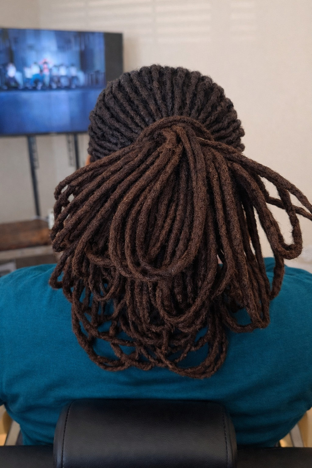 Person with medium brown dreadlocks sitting on a chair watching TV in a room with beige walls.