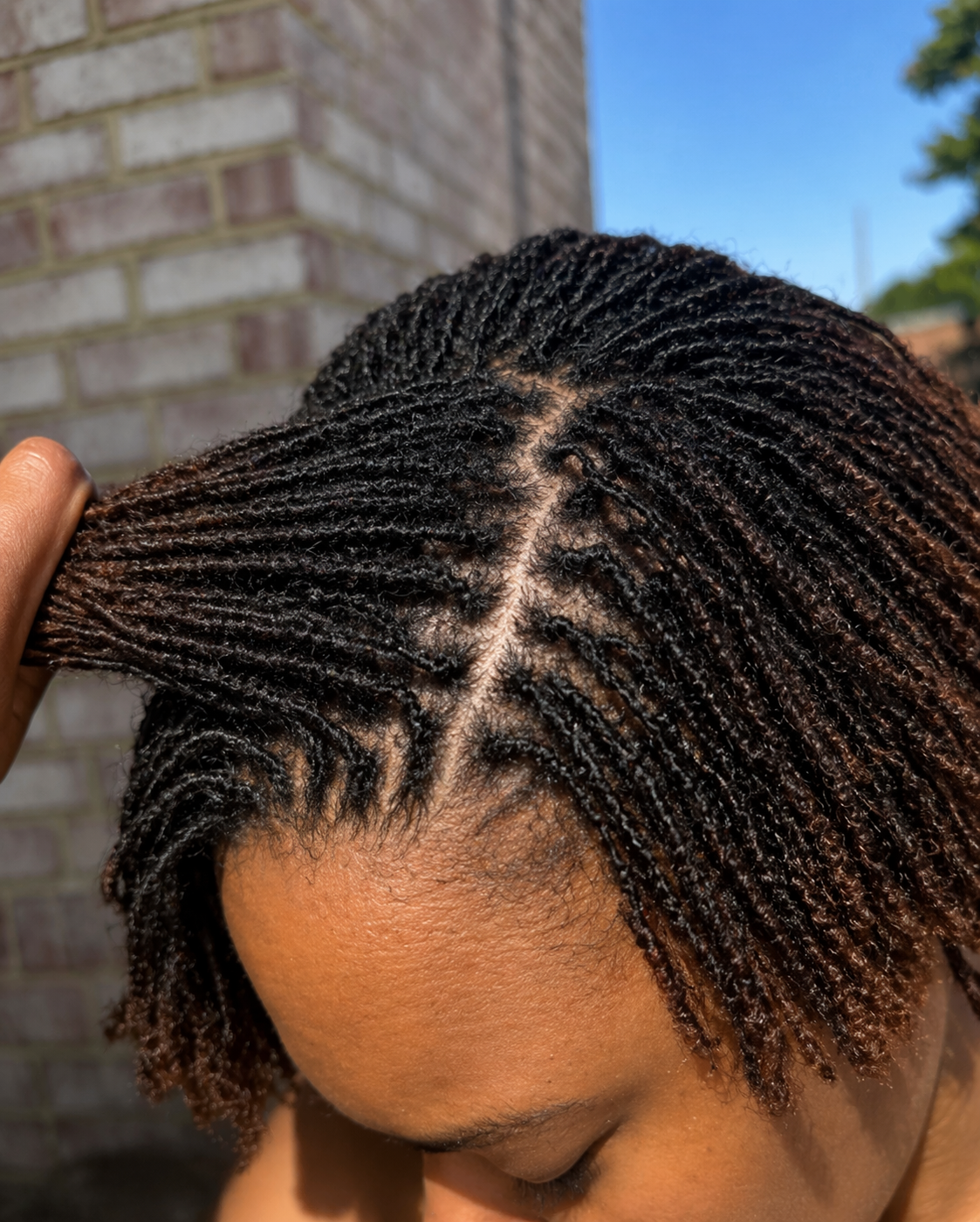 Close-up of a woman's head showing her freshly styled short twist braids with shiny, dark hair outdoors with a brick wall and blue sky in the background.