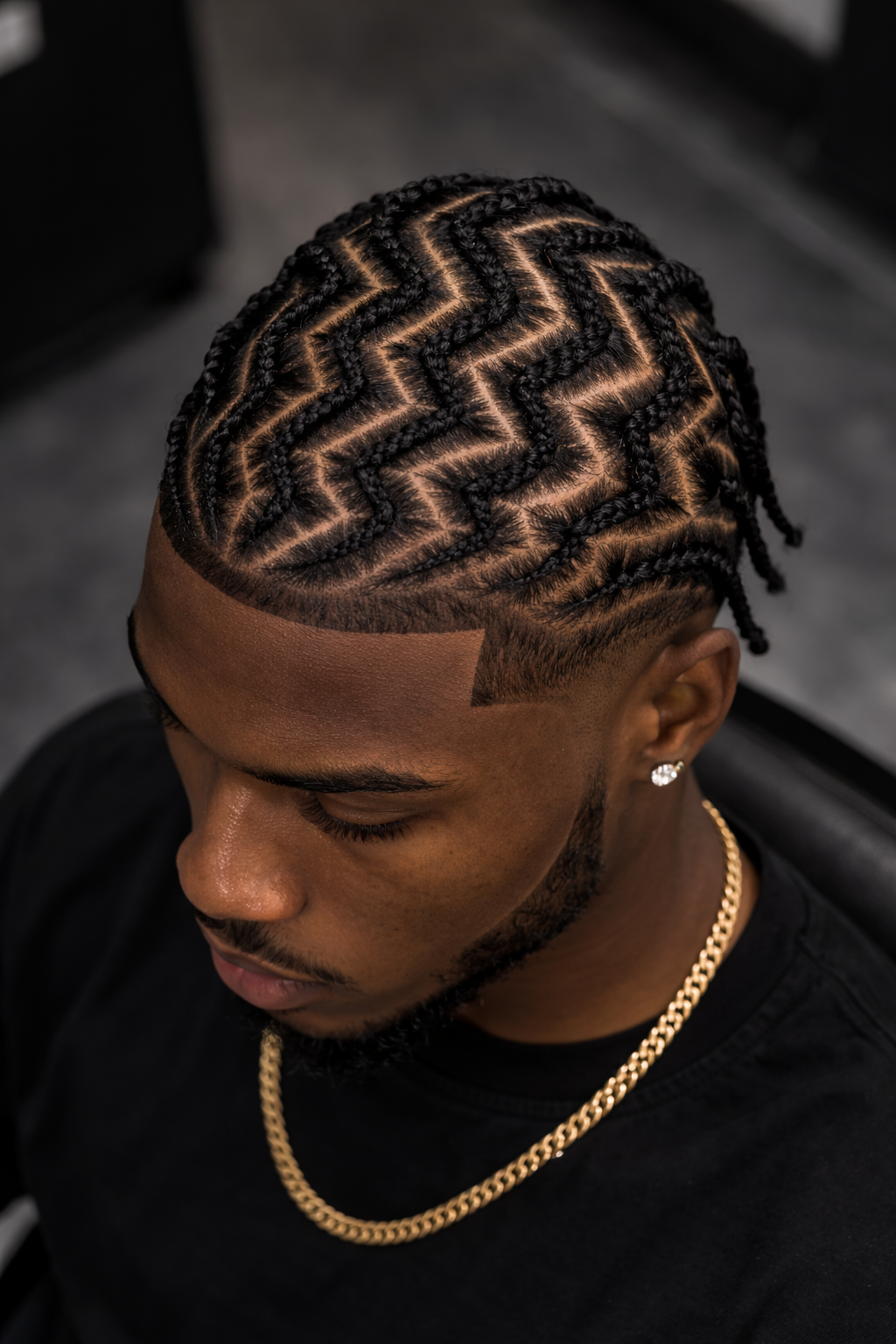 Close-up of a man with intricate braided hairstyle featuring a zigzag pattern, wearing a gold chain and earrings.