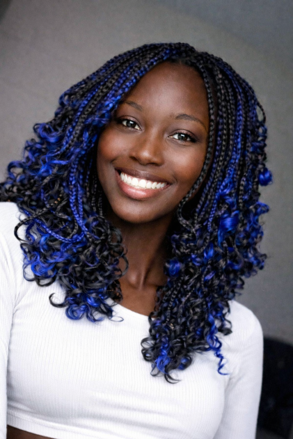 Portrait of a smiling woman with braided hair dyed blue and black, wearing a white top against a gray background.