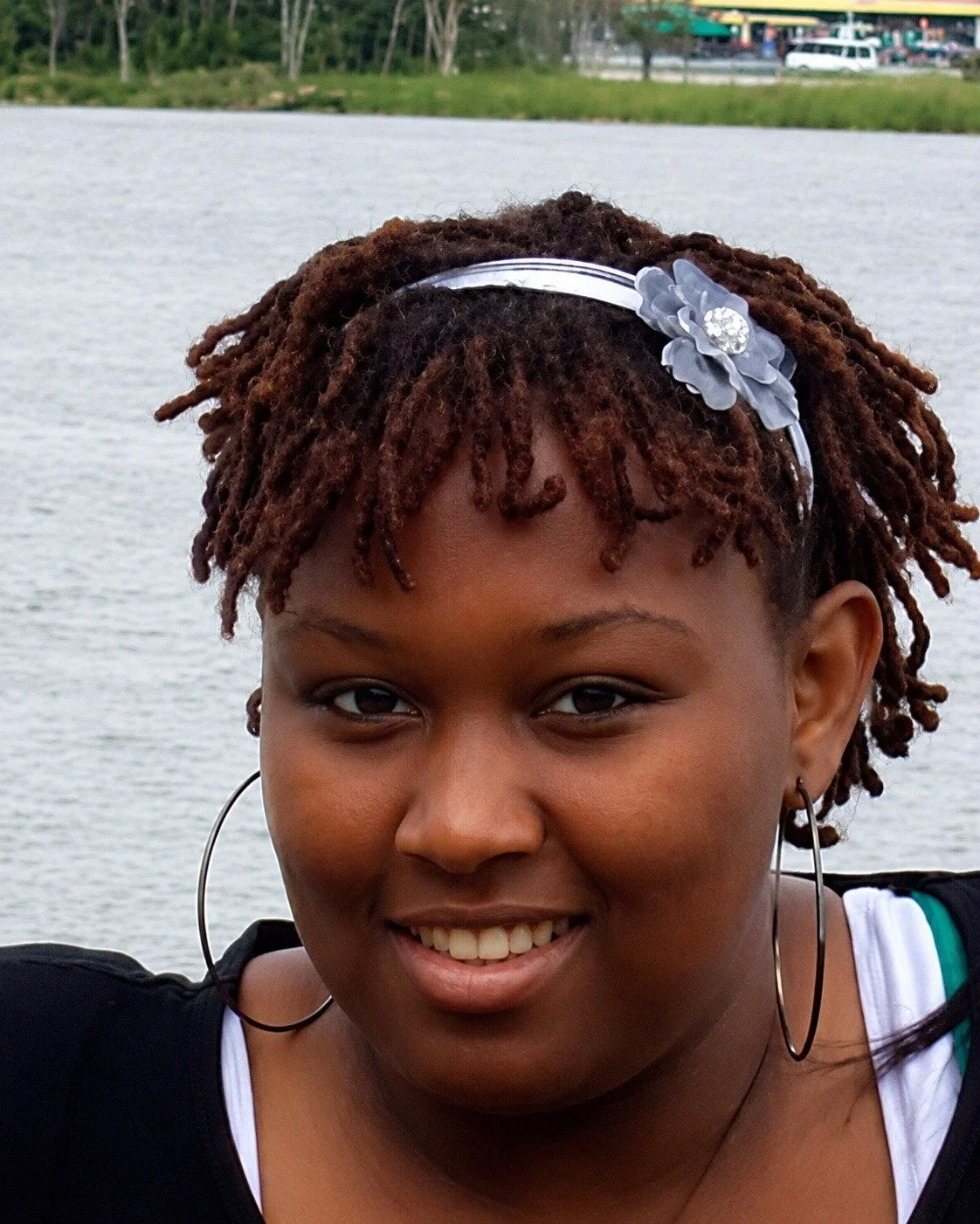 A portrait of a woman with short dreadlocks, wearing a black top, hoop earrings, and a floral headband, standing outdoors near a body of water with trees and buildings in the background.