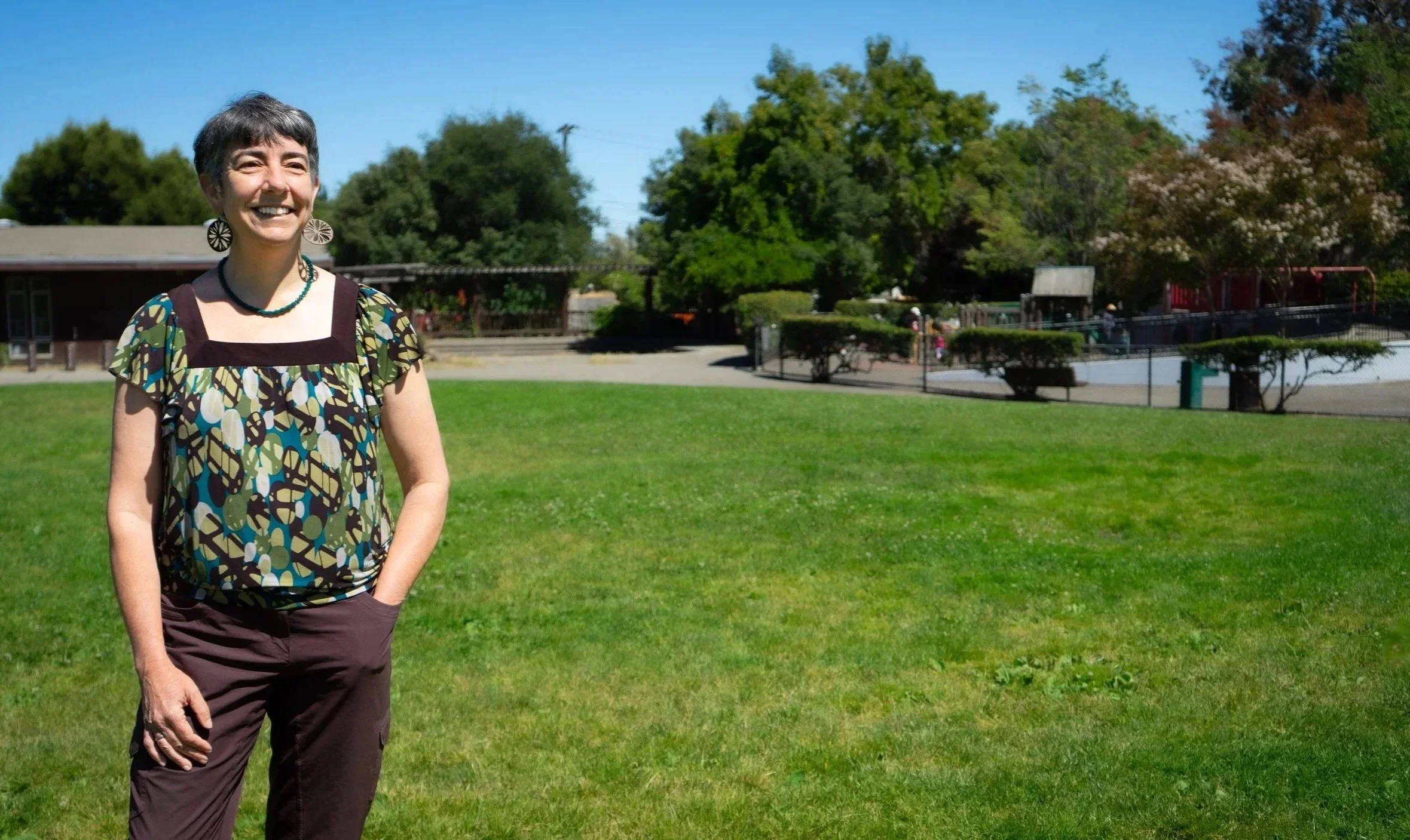 A woman with short dark hair and patterned earrings standing on a grassy field with trees and a playground in the background during sunny weather.