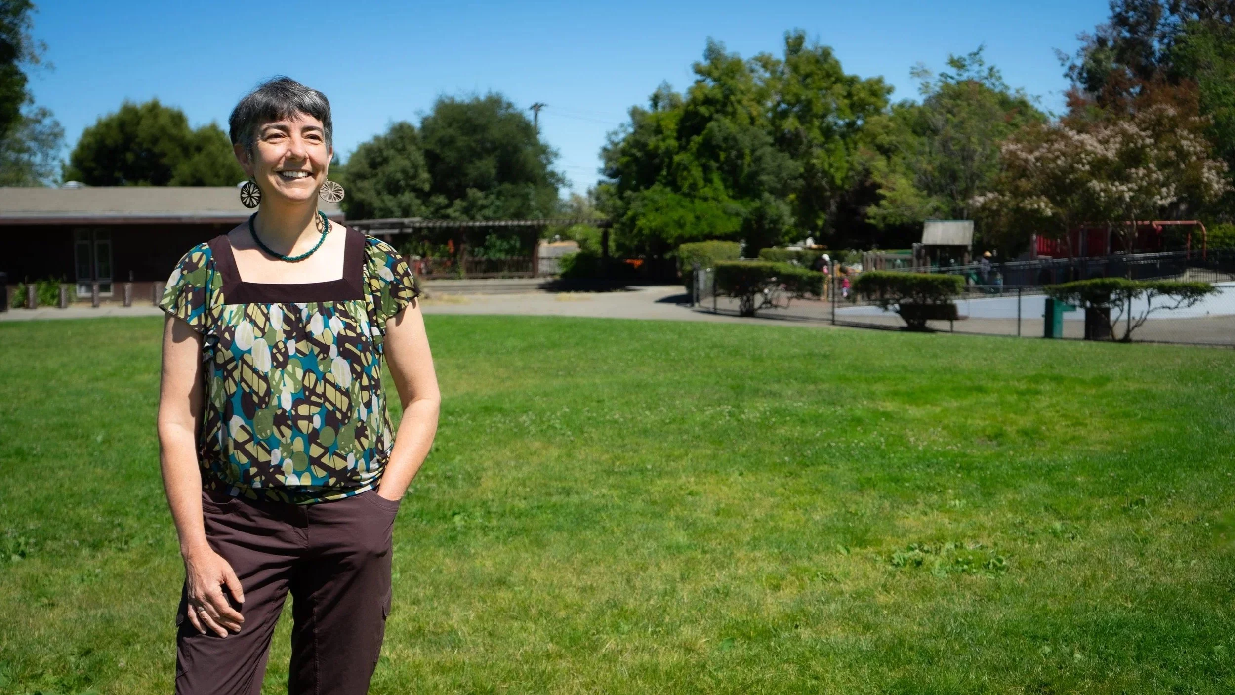 A woman standing outdoors on a sunny day with a big smile, wearing a colorful patterned top, earrings, and a necklace, with a grassy park and trees in the background.