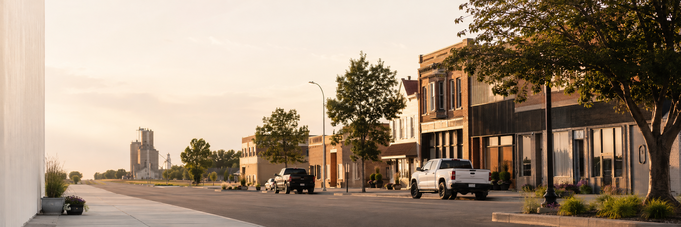 A quiet street lined with trees and brick buildings at sunset, with parked cars and industrial structures in the distance.
