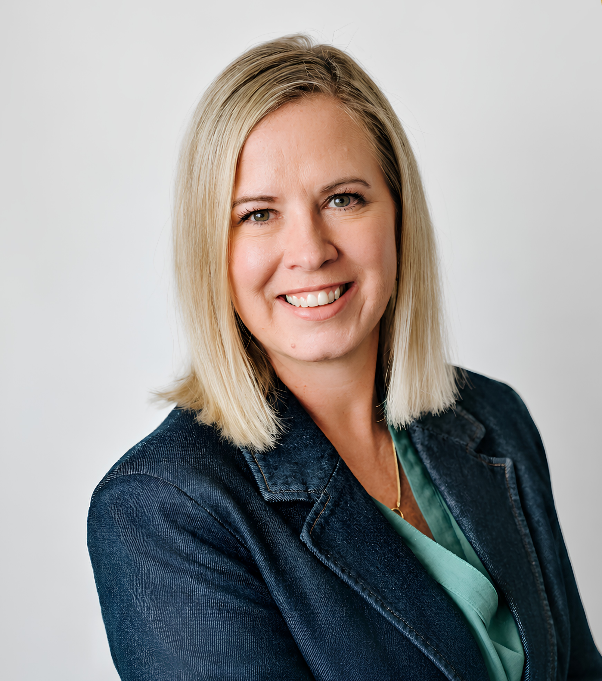 A smiling woman with blonde hair wearing a dark jacket and a green blouse, standing against a plain white background.