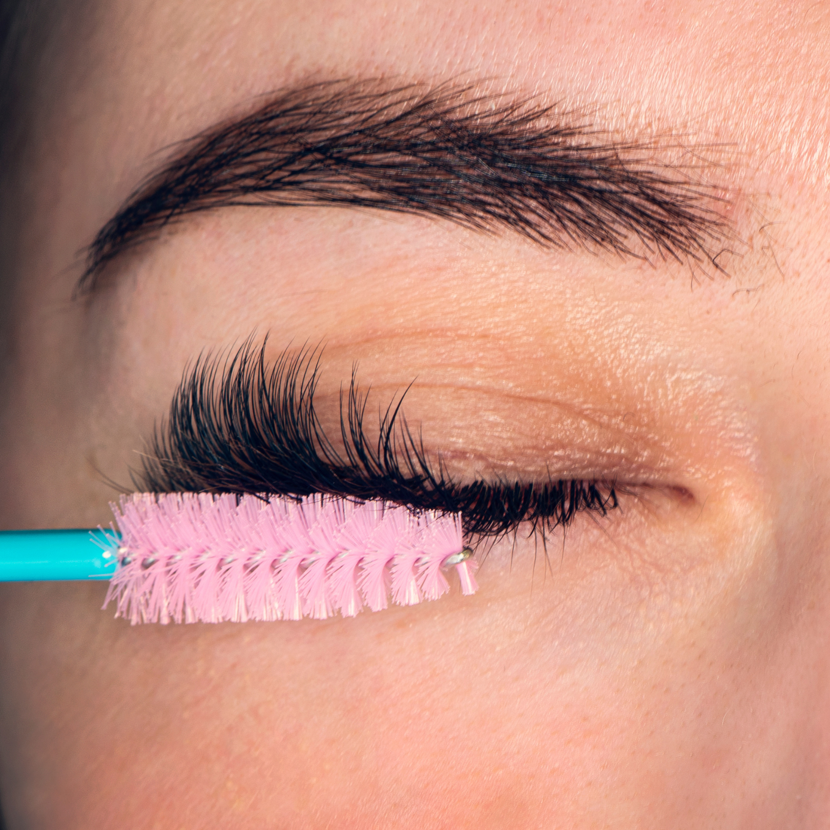 Close-up of a closed eye with makeup, being cleaned or prepared with a pink eyelash brush.