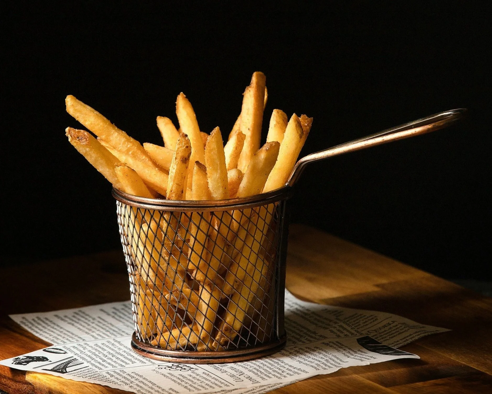 Fried French fries in a metal wire basket on a wooden table with a black background.