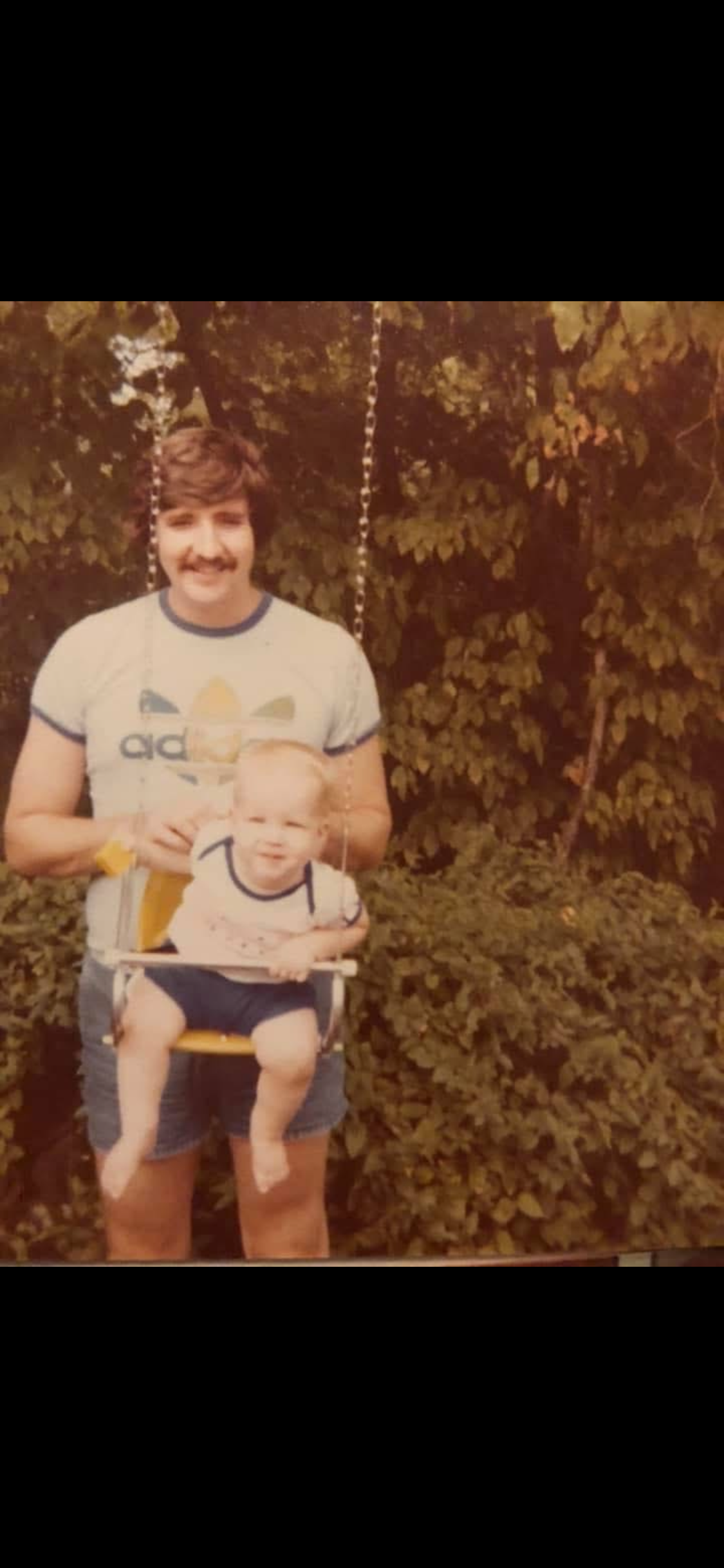 A young man with brown hair and a mustache standing outdoors in front of bushes, holding a small child in a high chair. The young man is wearing a vintage Adidas t-shirt and shorts, smiling at the camera. The child has light hair and is wearing a striped shirt, sitting in a yellow high chair with a tray, looking slightly to the side.