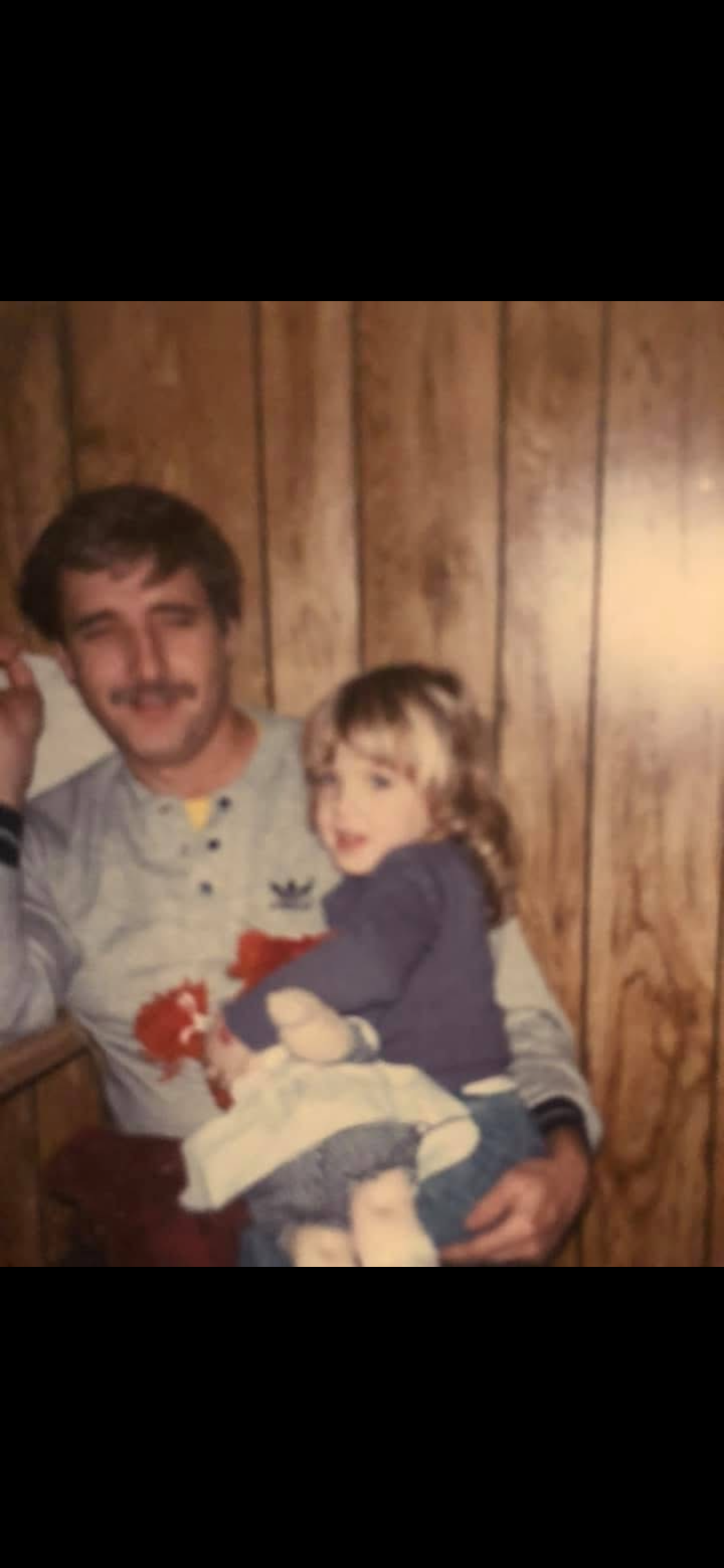 A man with dark hair and a mustache holding a young girl, both smiling, sitting against a wood-paneled wall. The girl is holding a cake with a snowman decoration, and she has light brown hair and is wearing a dark shirt.