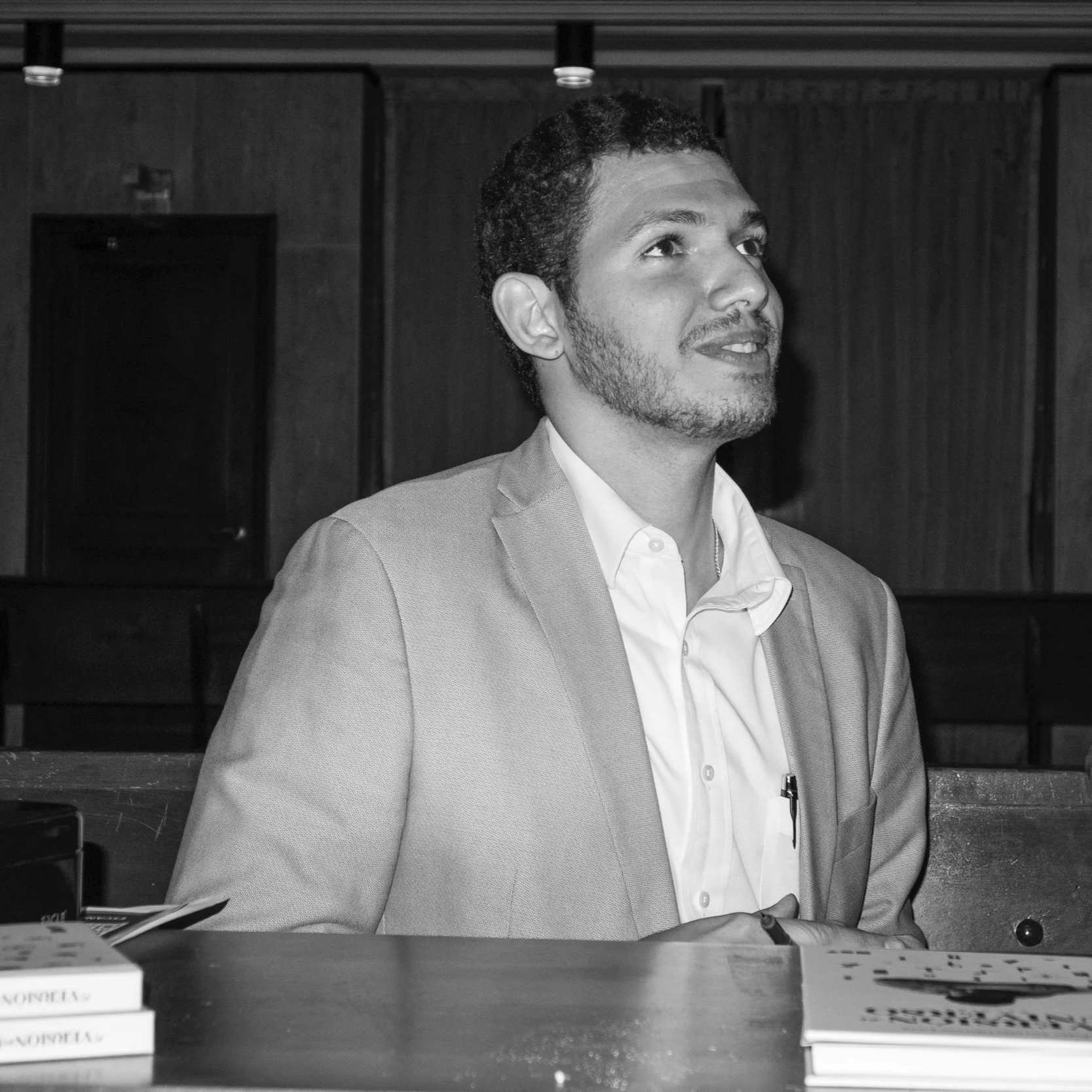 Escritor con barba y cabello rizado, vestido con un saco gris y camisa blanca, sentado en un entorno interior en la puesta en circulación de su nuevo libro.