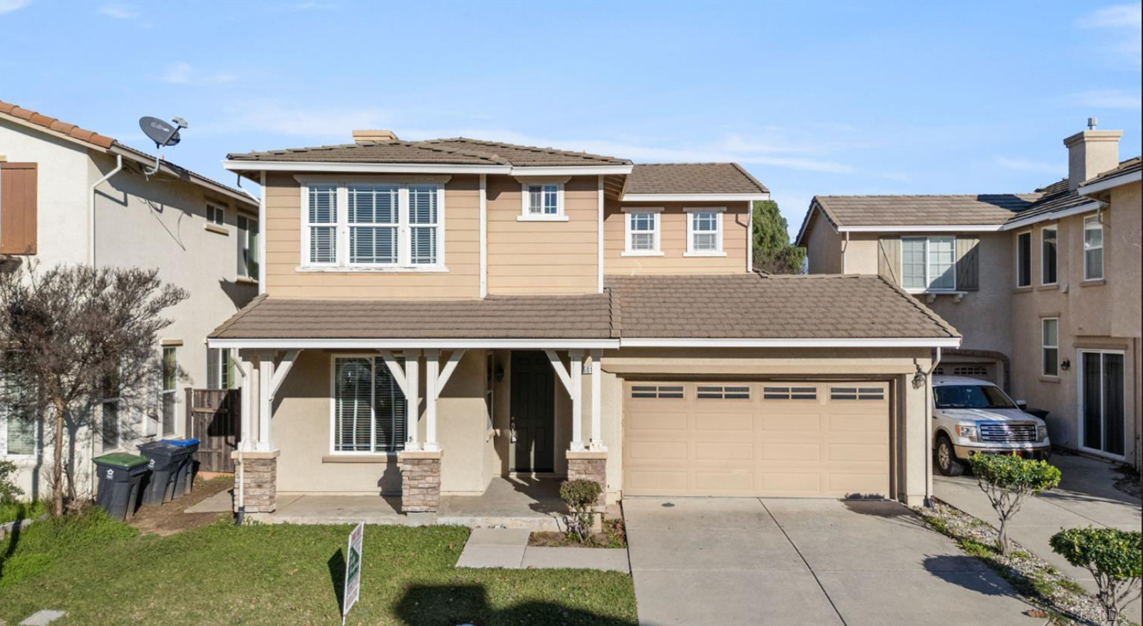 Two-story suburban house with beige siding, a front porch, a garage, and a small front yard with trees and shrubs.