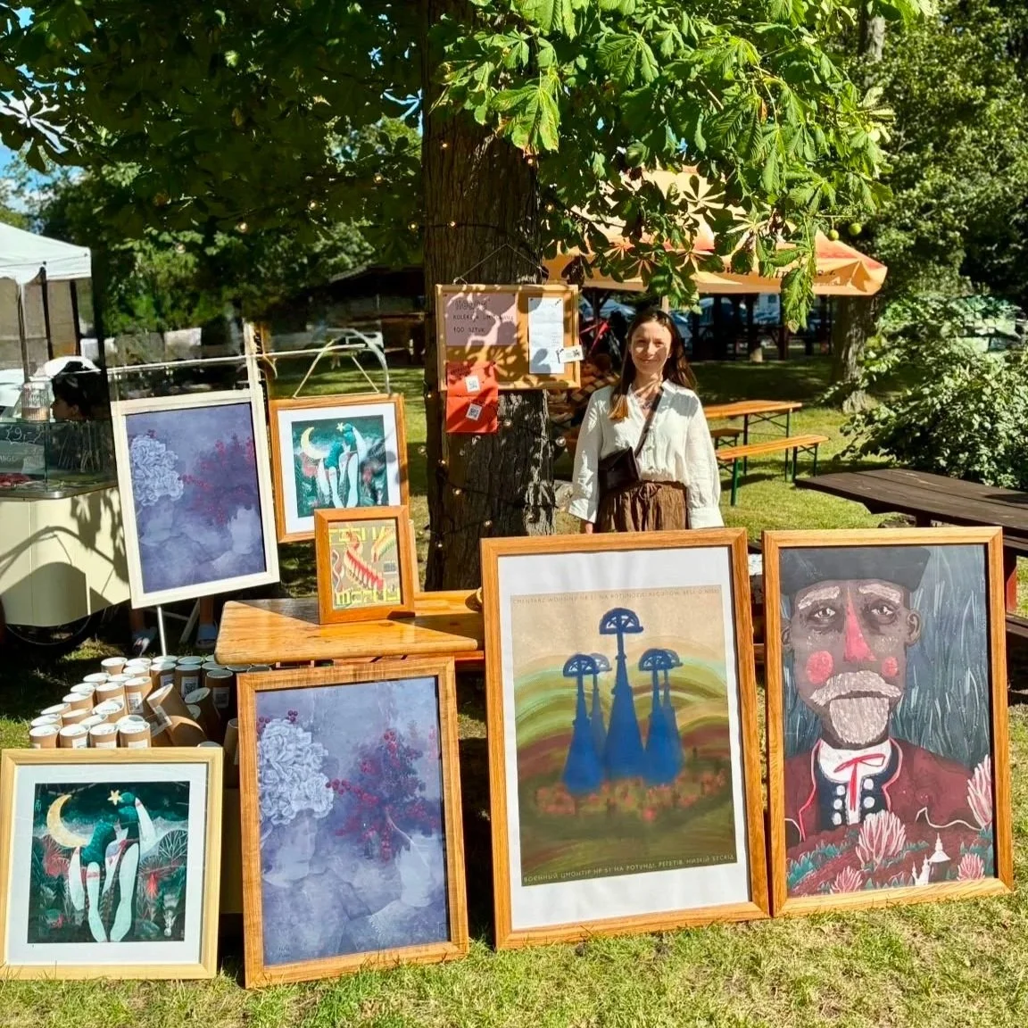 Łemkowska artystka przy swoim stoisku podczas plenerowego jarmarku sztuki, otoczona plakatami i obrazami prezentowanymi w naturalnych drewnianych ramach z rodzinnej produkcji.