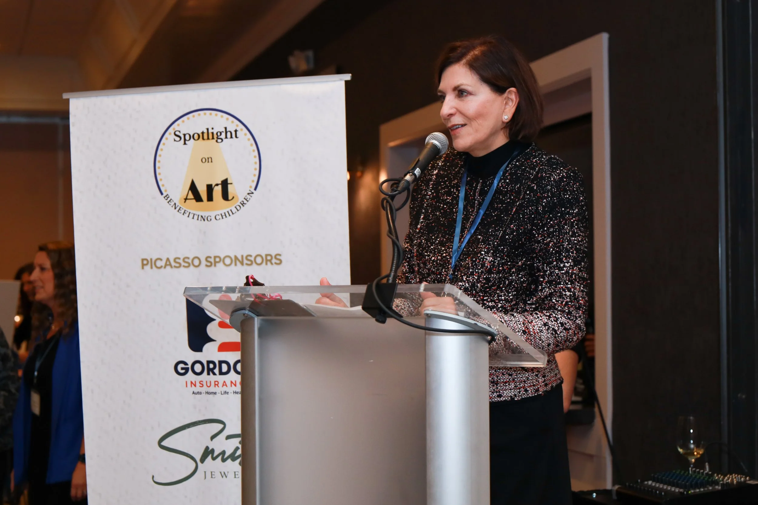 A woman with short brown hair speaking into a microphone at a podium during an event. A banner in front of her displays the text "Spotlight on Art benefiting children" and sponsors, with additional logos and text.