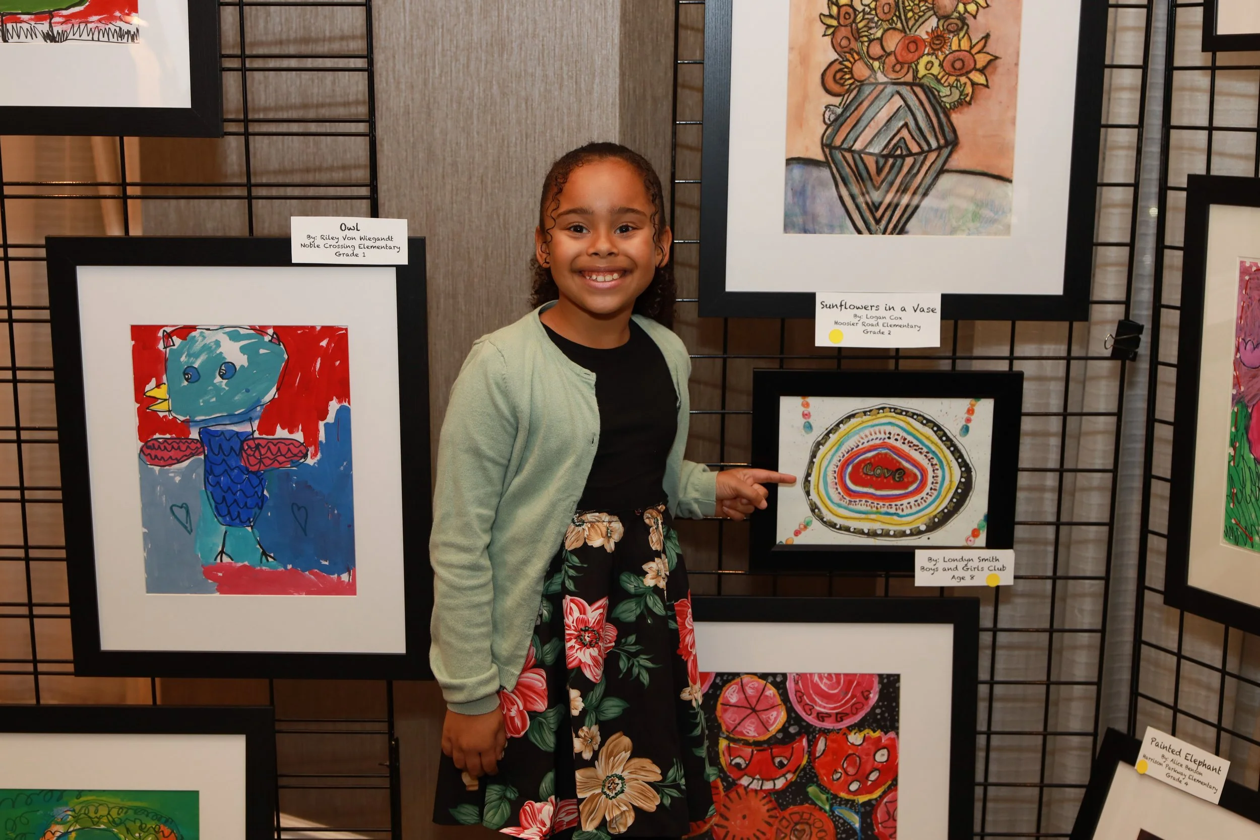 Young girl at an art display, smiling, wearing a black shirt, floral skirt, and light green cardigan. Artwork behind her includes colorful paintings and drawings, some depicting animals and abstract designs.