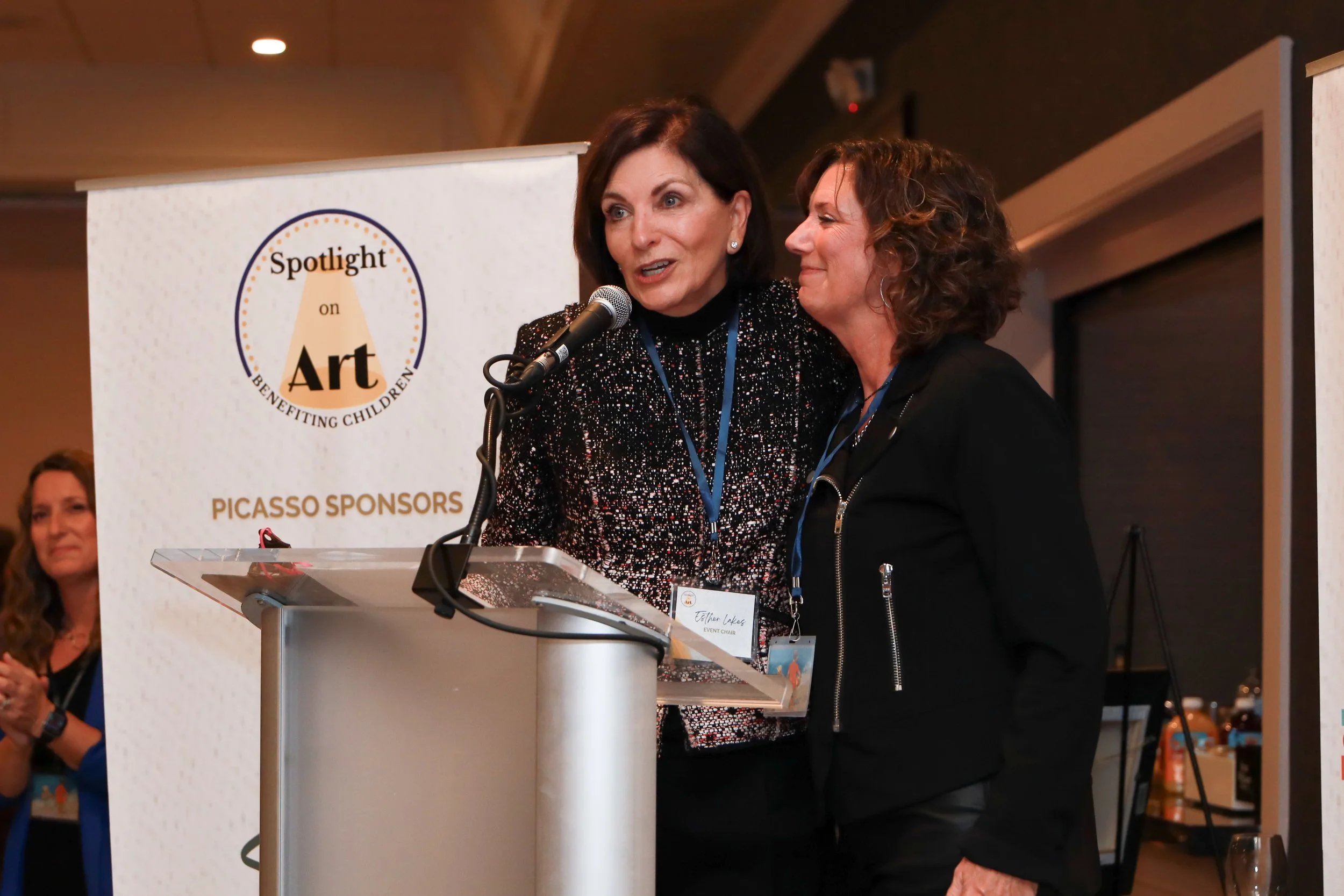 Two women standing at a podium during an event, with one speaking into a microphone and the other smiling. Behind them is a banner that reads "Spotlight on Art - Benefiting Children".