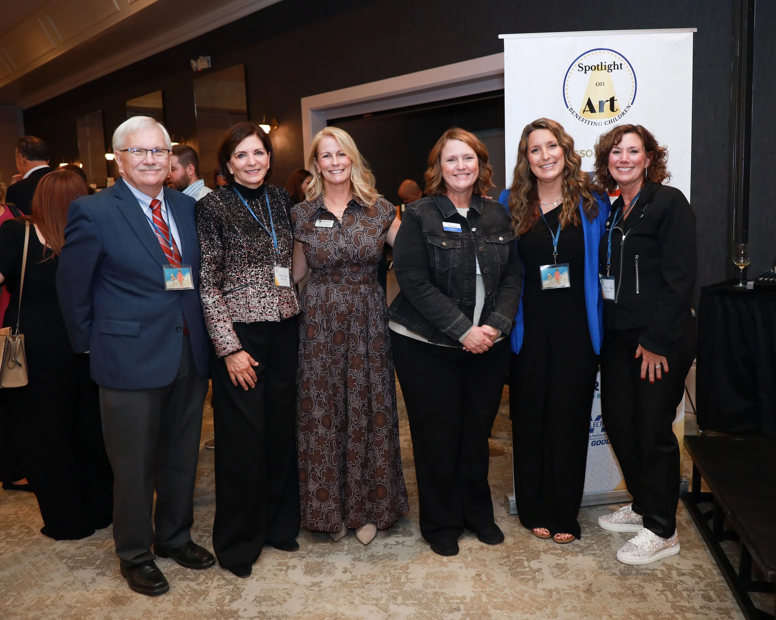 Group of six women and one man standing together at an indoor event, smiling for the camera, with a banner behind them reading 'Spotlight on Art Benefiting Children'.