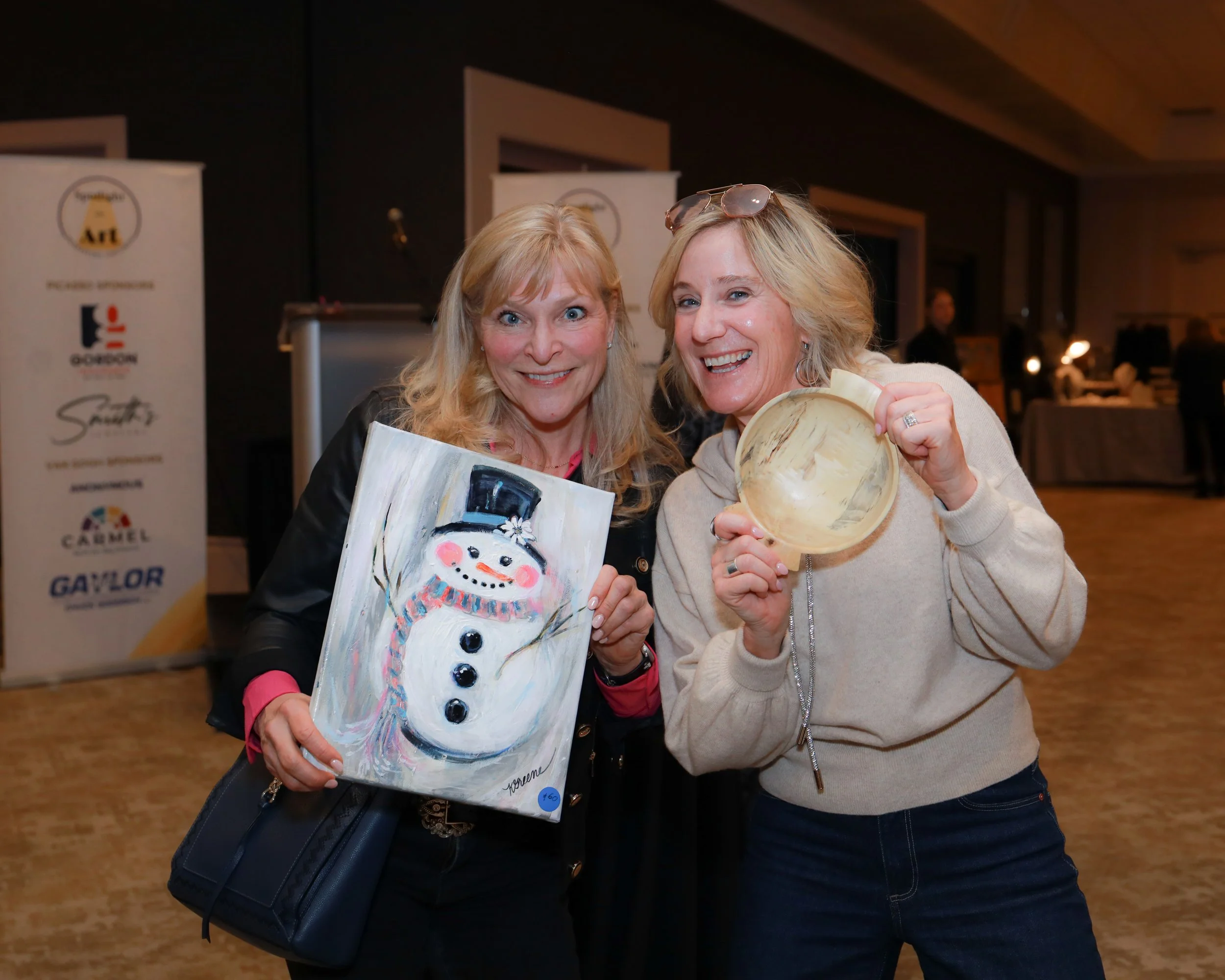 Two women smiling and holding paintings at an art event, with banners and other people in the background.